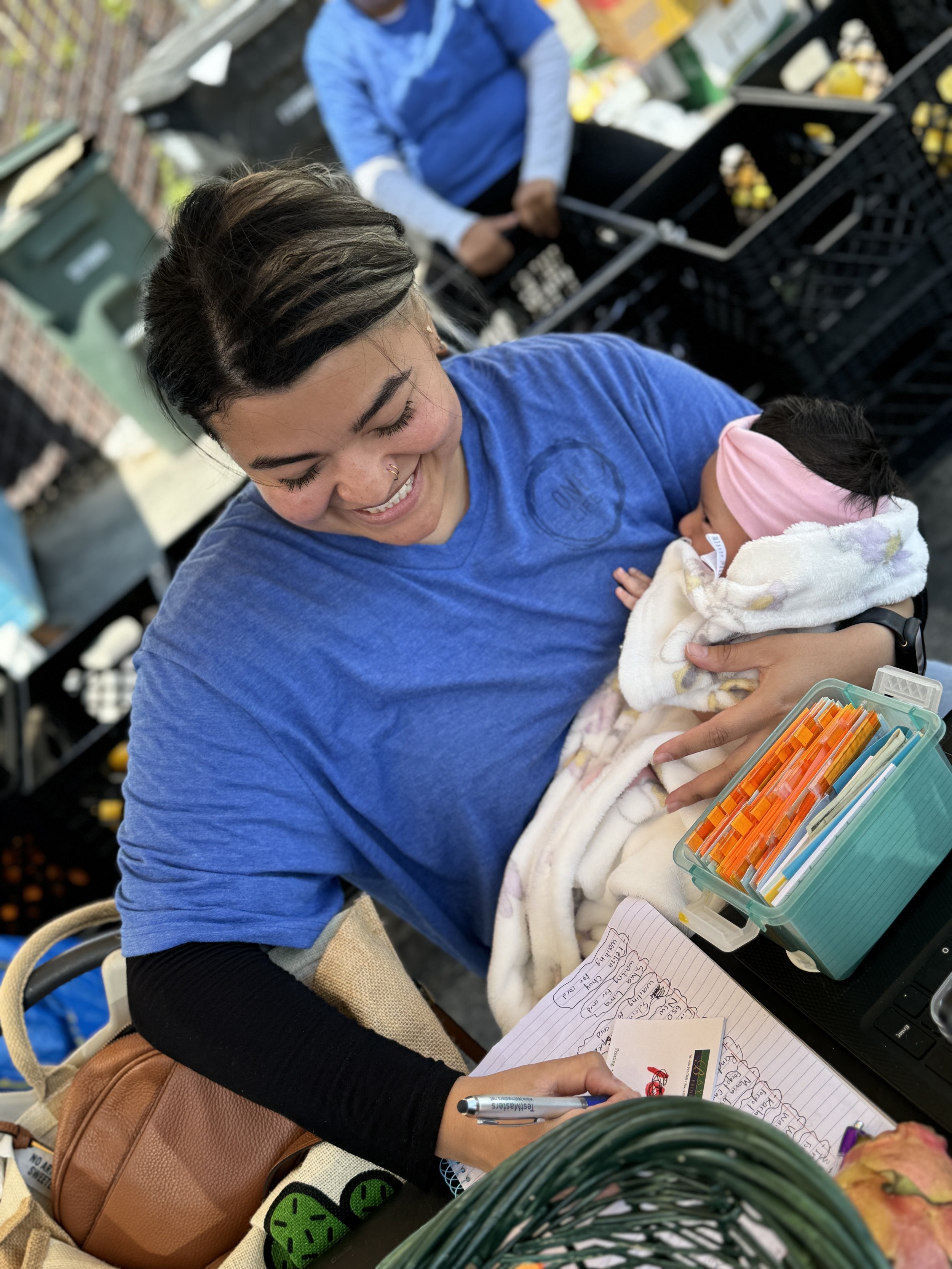 A woman holding a baby and writing notes at a table in an indoor store or market, surrounded by boxes of produce and shopping items.