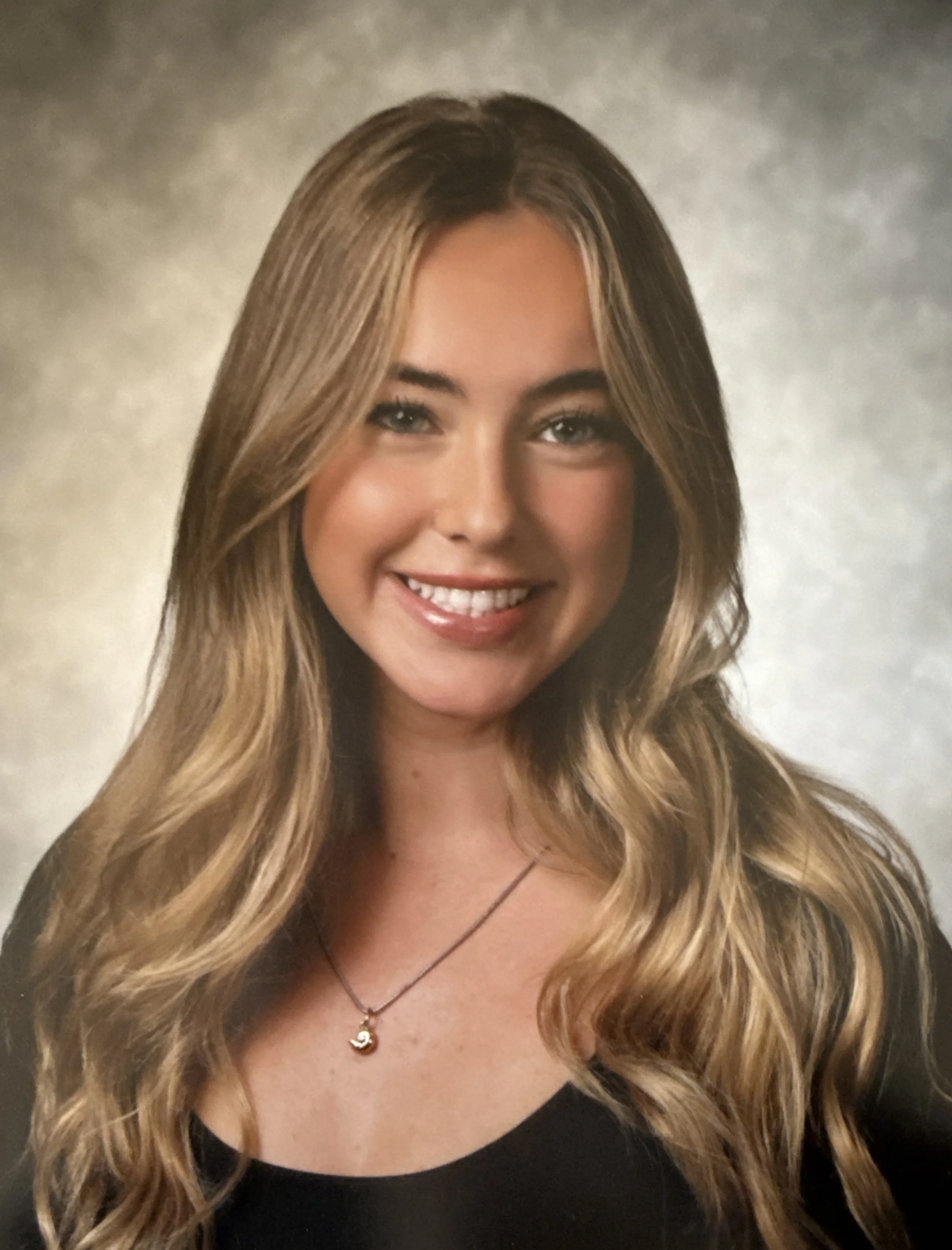Close-up portrait of a young woman with long, wavy brown hair, wearing a patterned top and layered necklaces, standing in front of a wooden fence.