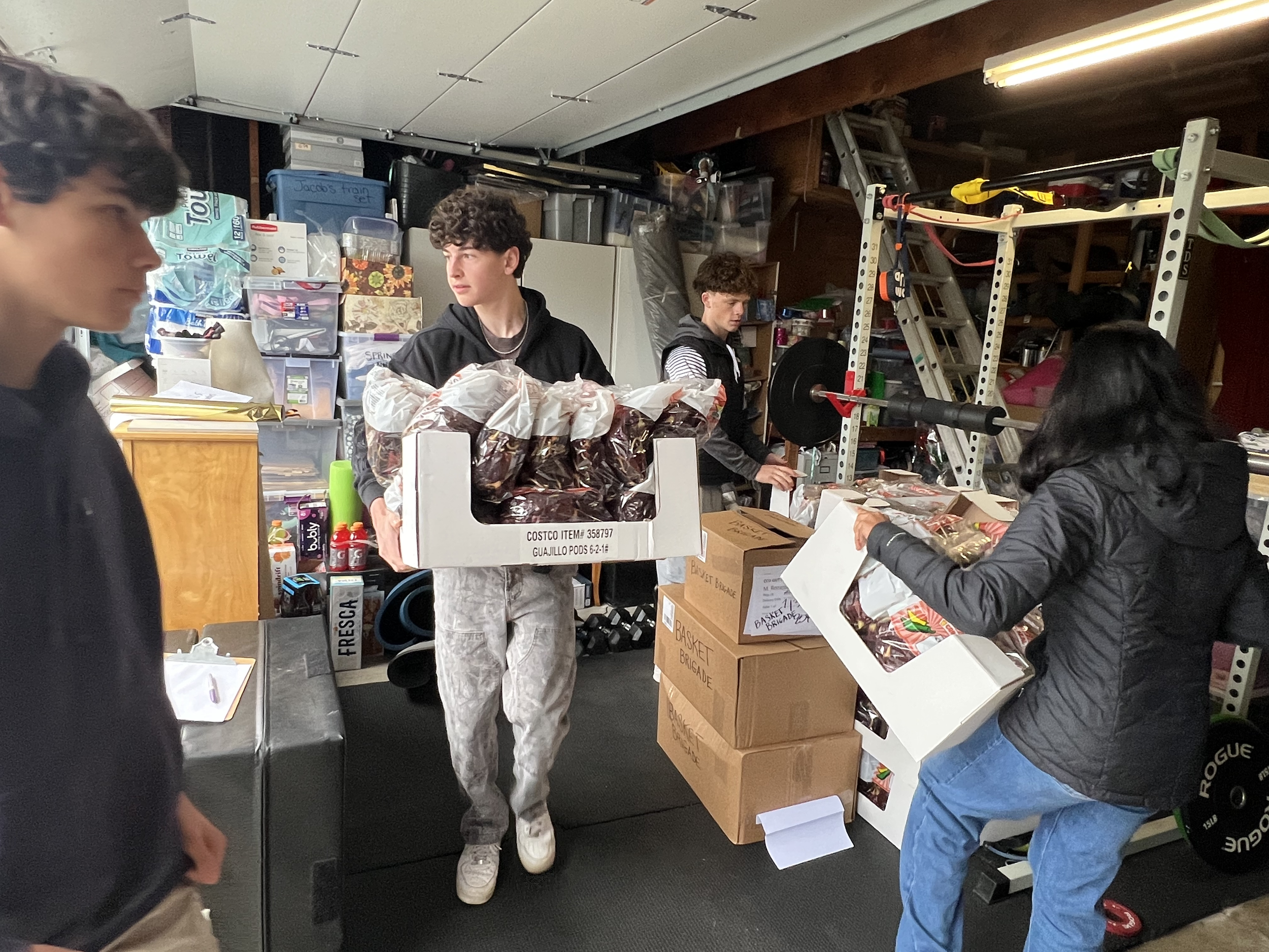 Four people organizing packages in a garage or storage room with shelves, boxes, and workout equipment.