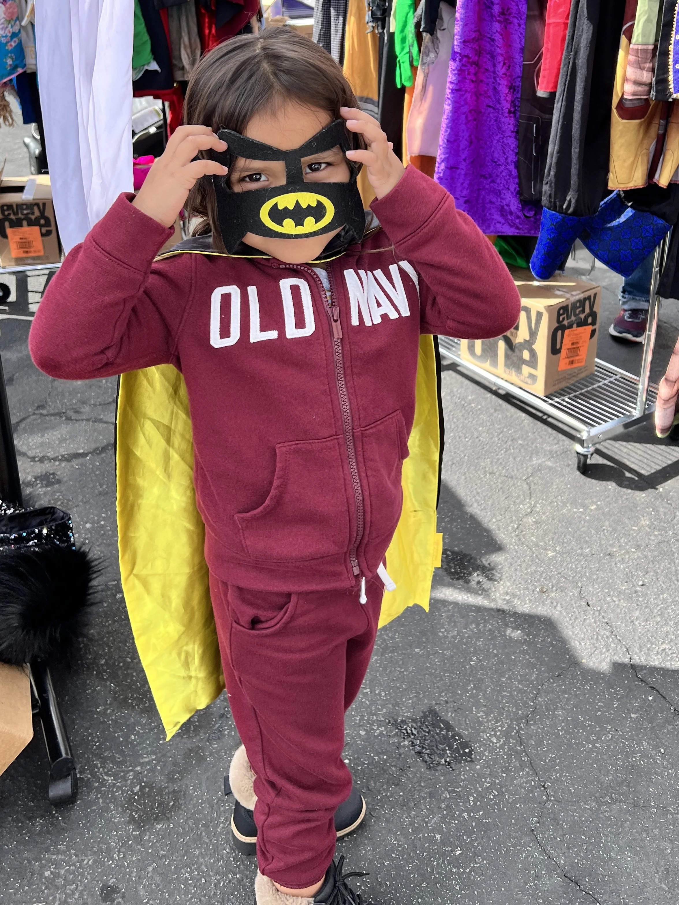 A young girl wearing a Batman mask, a red hoodie with "Old Navy" written on it, and red pants, standing outdoors at a market or fair. She has a yellow cape and is adjusting her mask with a background of hanging clothing and boxes.