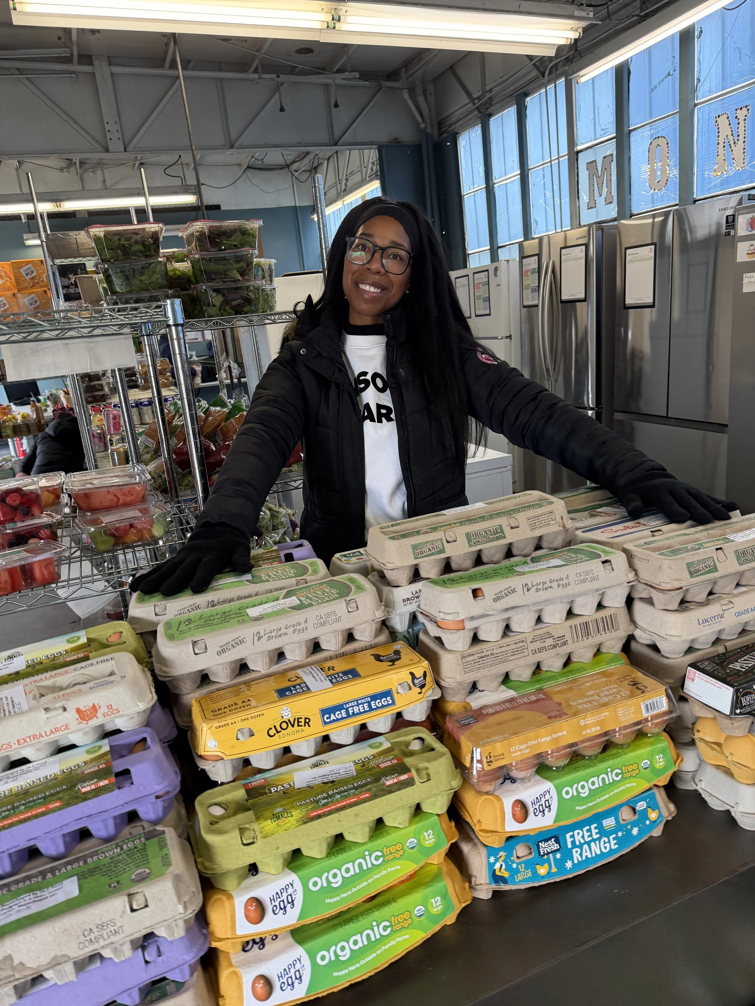 A woman wearing glasses, a black jacket, and black gloves smiling behind a display of eggs at a grocery store or farmers market.
