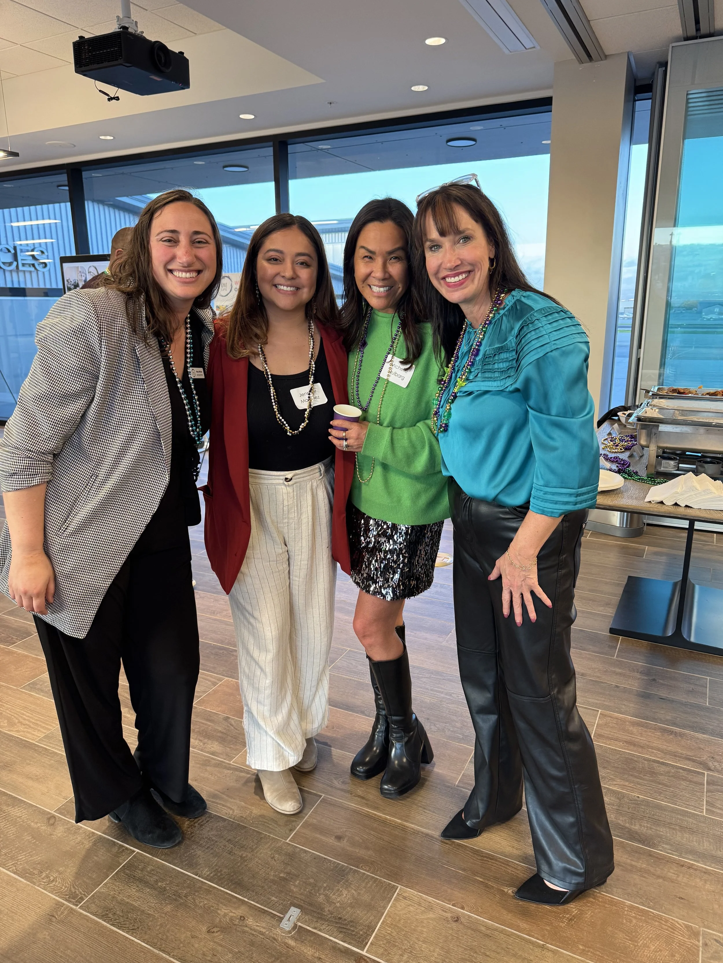 Four women smiling and posing for a photo at a social event with name tags, beads, and drinks, standing on a wooden floor near large windows.