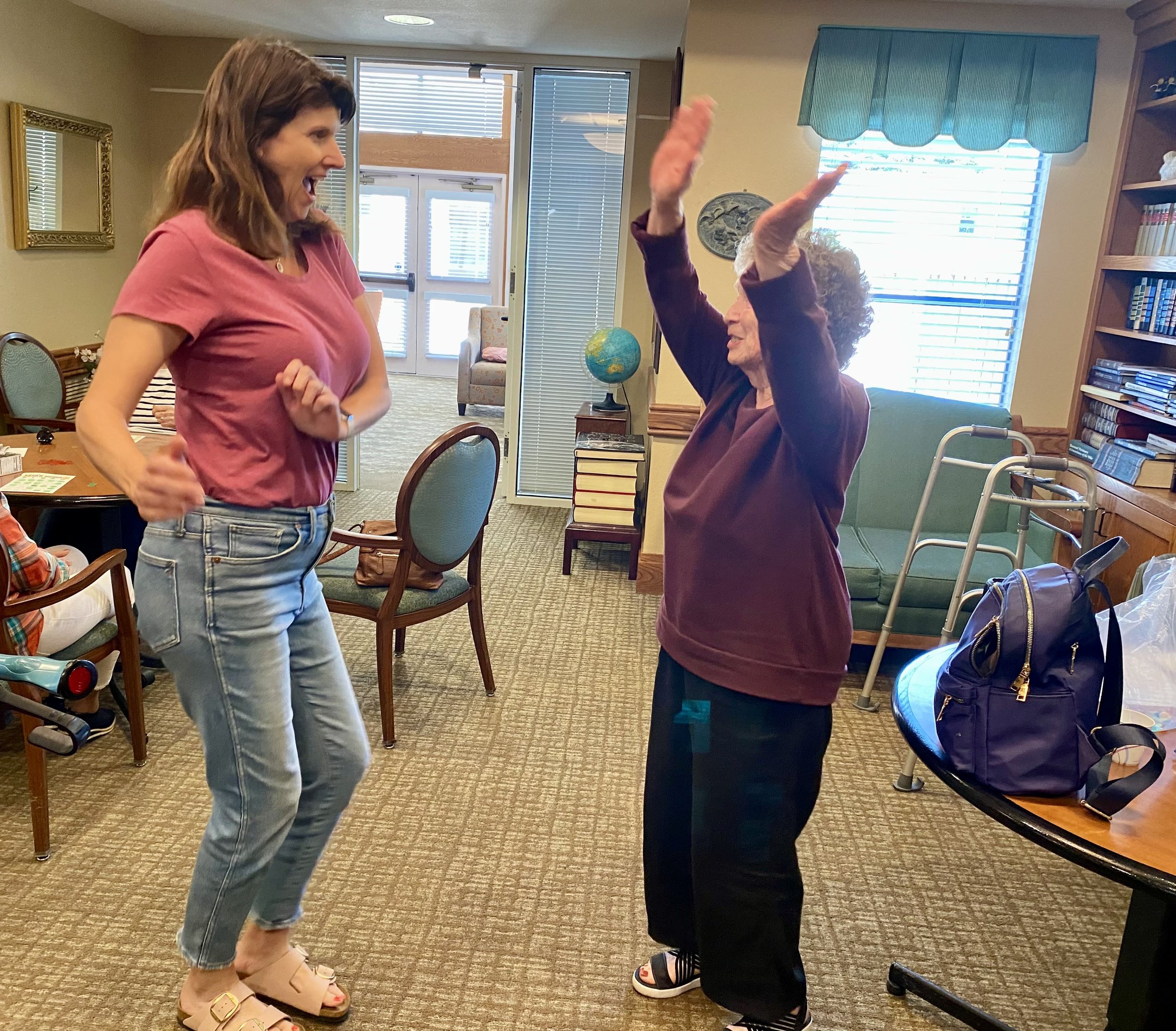 Two women are dancing and smiling indoors, with one woman raising her hands. The scene appears joyful and lively, set in a room with chairs, a bookshelf, and a table.