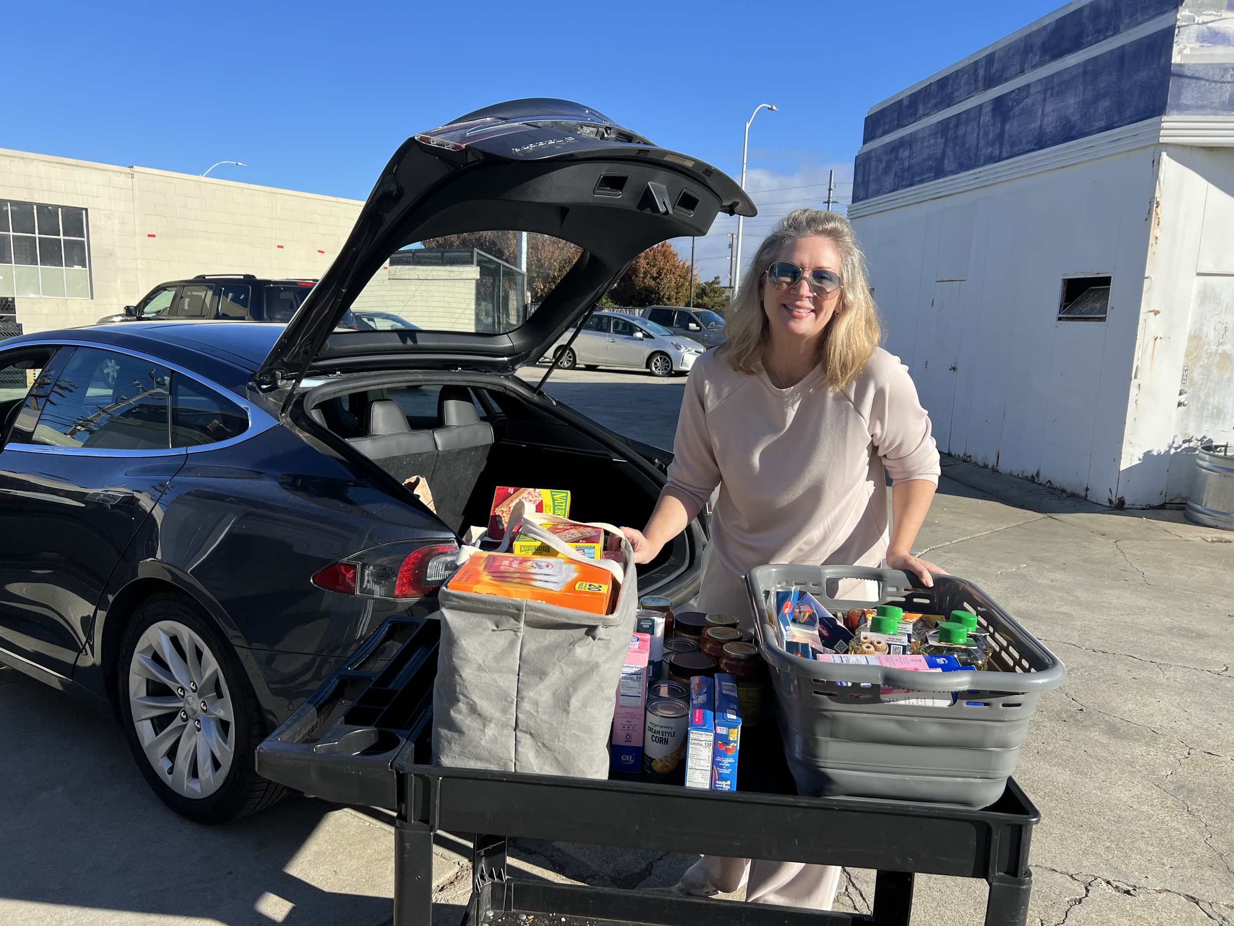 A woman with blonde hair wearing sunglasses and a beige sweater loading groceries into the trunk and shopping cart of a black electric car parked outdoors in a commercial area on a sunny day.