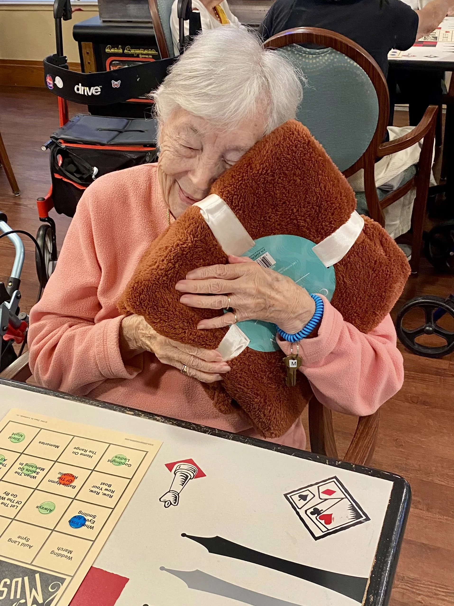 An elderly woman with white hair hugging a large stuffed teddy bear at a table in a social setting, smiling and holding the bear close.