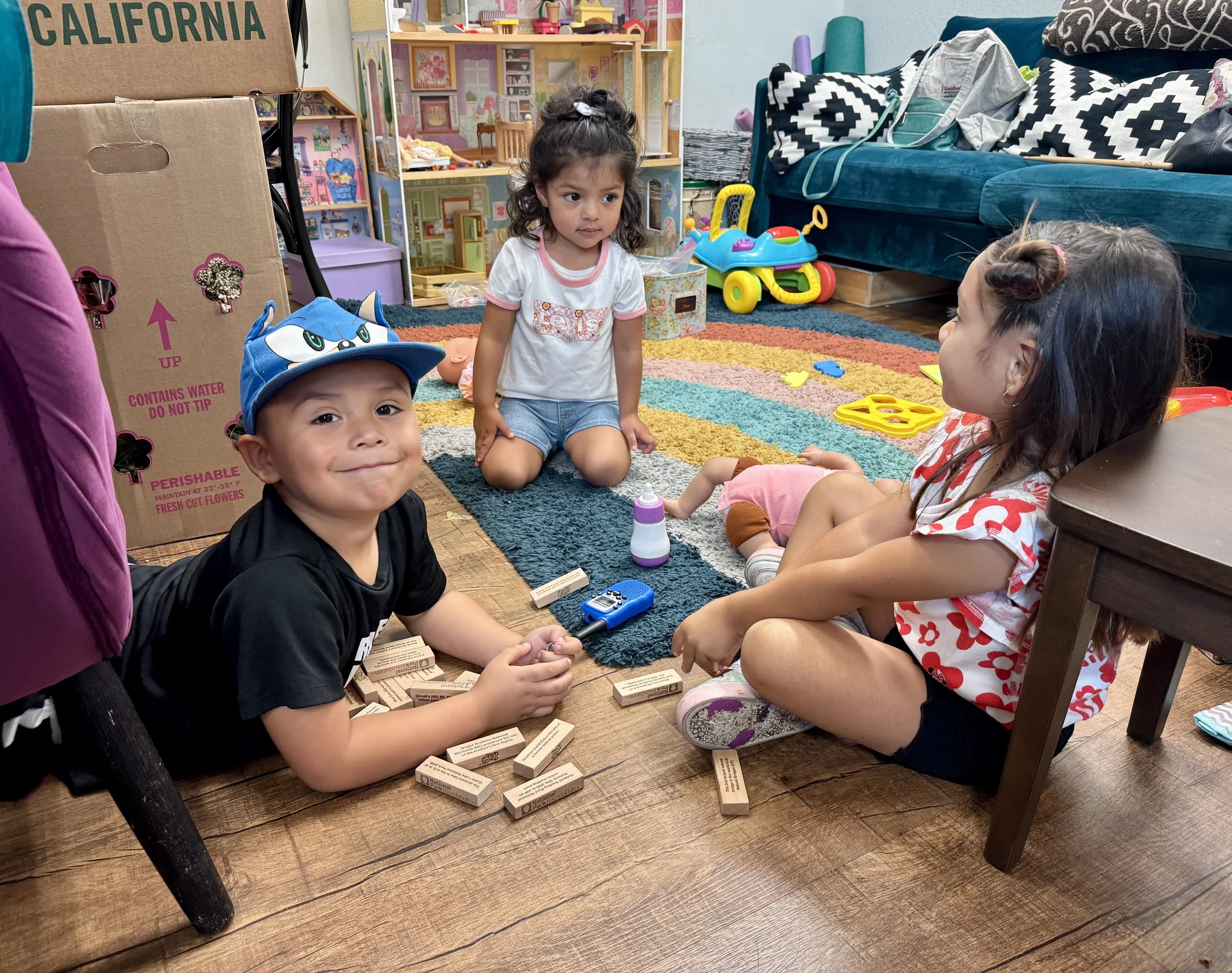 Three children playing on a wooden floor in a playroom, surrounded by toys, including wooden blocks, a baby doll, and a toy bottle. One boy in a Sonic the Hedgehog hat smiles at the camera, while two girls sit nearby, one with a bun hairstyle and a c