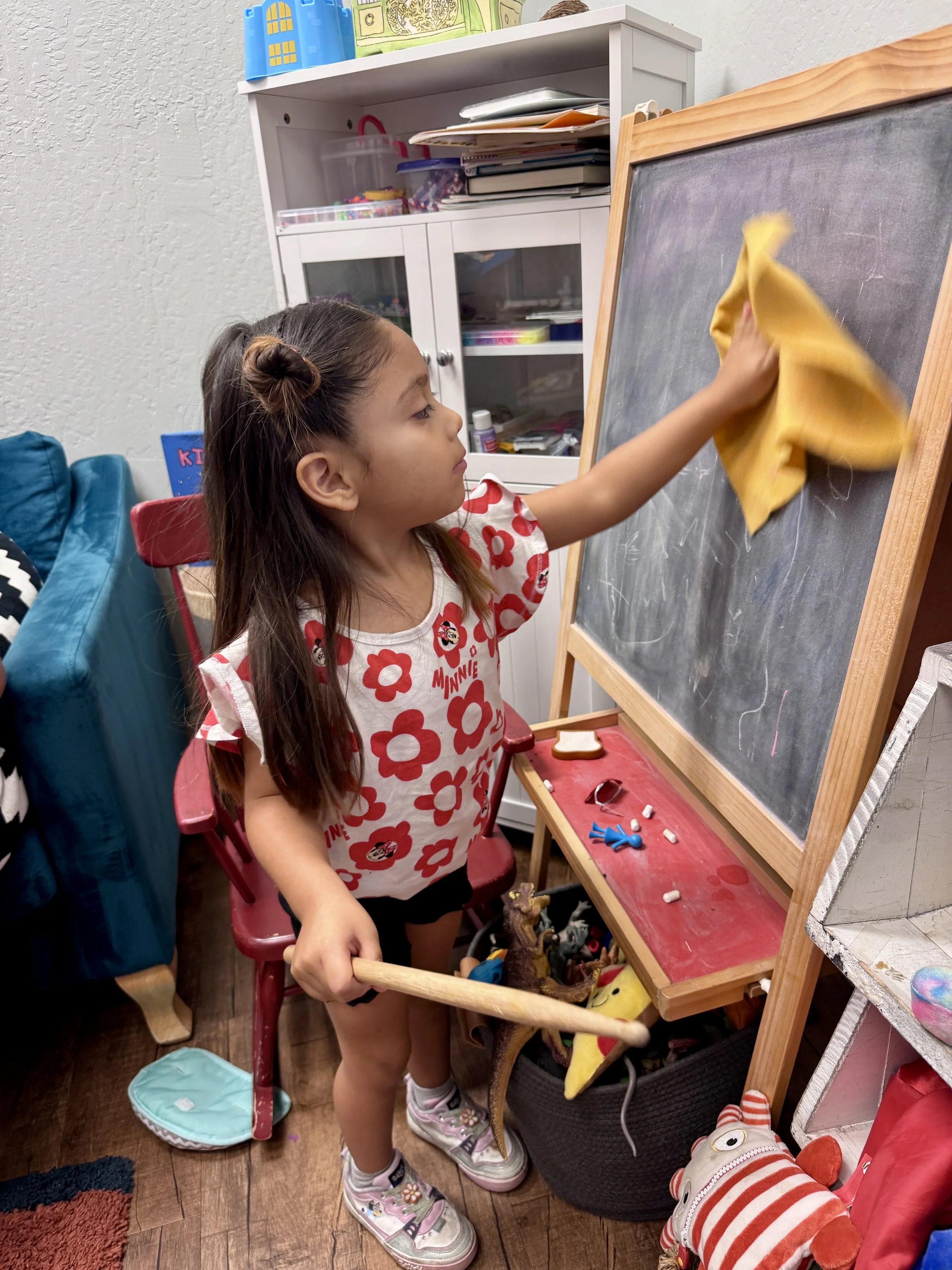 A young girl with a bun hairstyle, wearing a Minnie Mouse shirt, is cleaning a small chalkboard with a yellow cloth. She stands on a red chair, holding a wooden stick. The room has toys on the floor, a white cabinet with books and supplies, and a plu