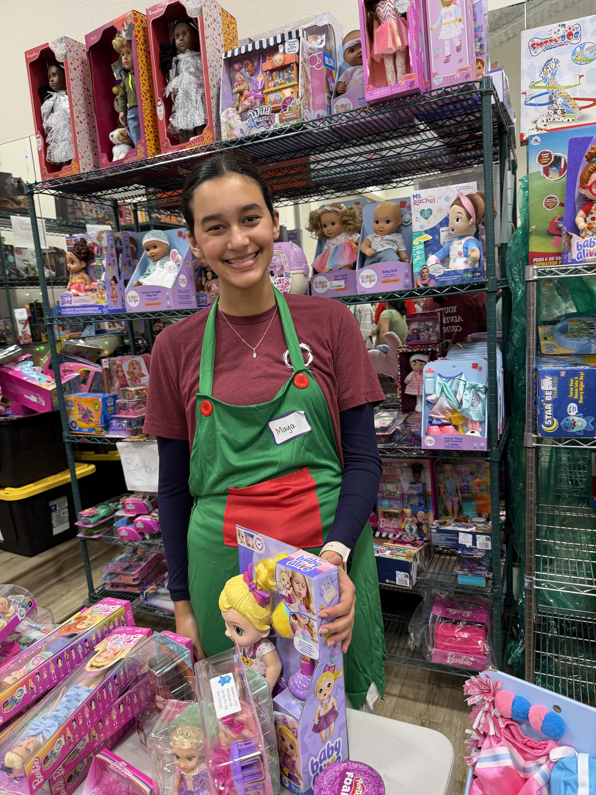 A smiling woman wearing a green apron with a name tag that says 'Maya' stands behind a table filled with dolls and toys in a toy store. Shelves behind her are stocked with various dolls and children's toys in colorful packaging.
