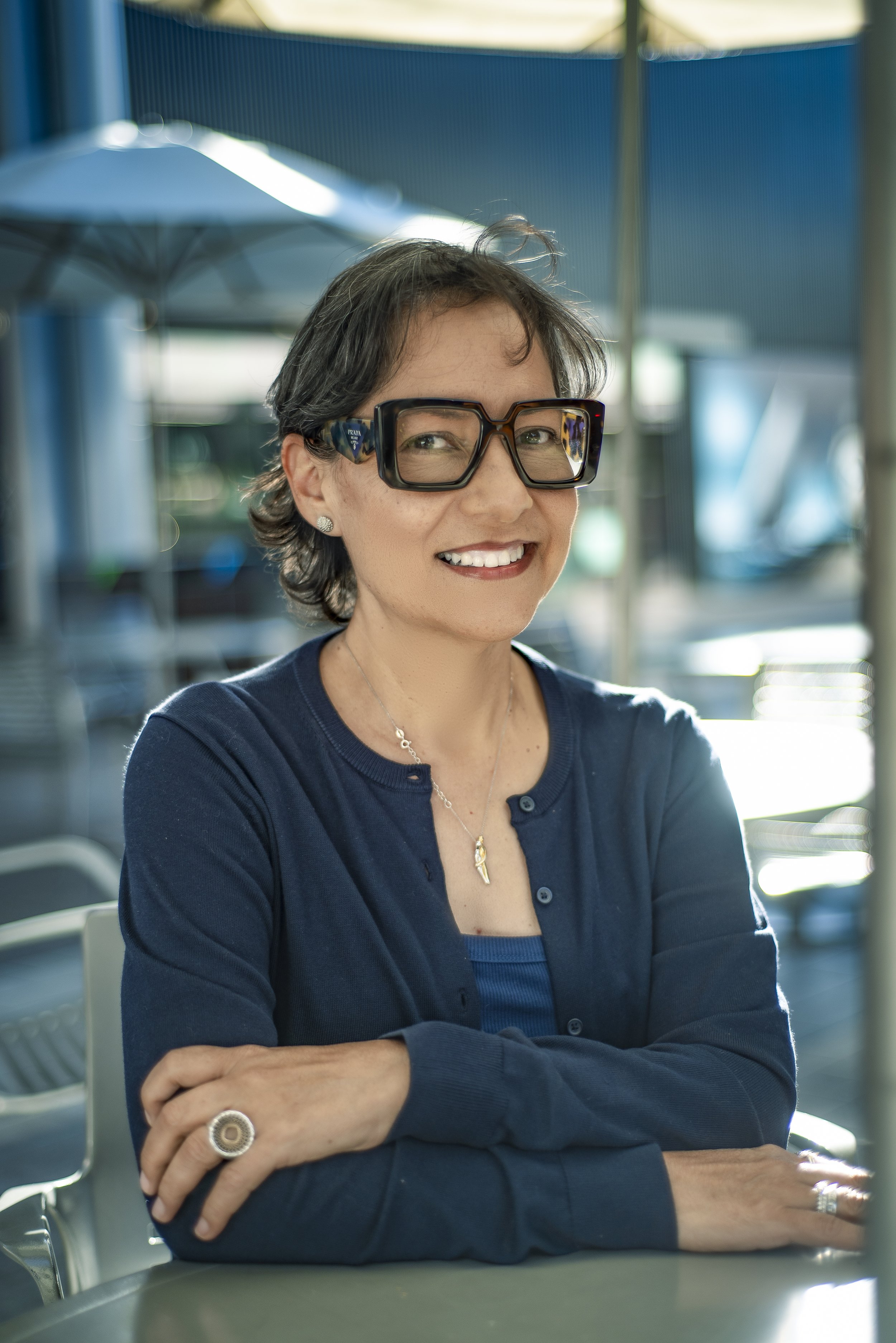 A woman with short dark hair, wearing large glasses, smiling, sitting at a table outdoors with blue umbrellas and a blue fence or wall in the background.