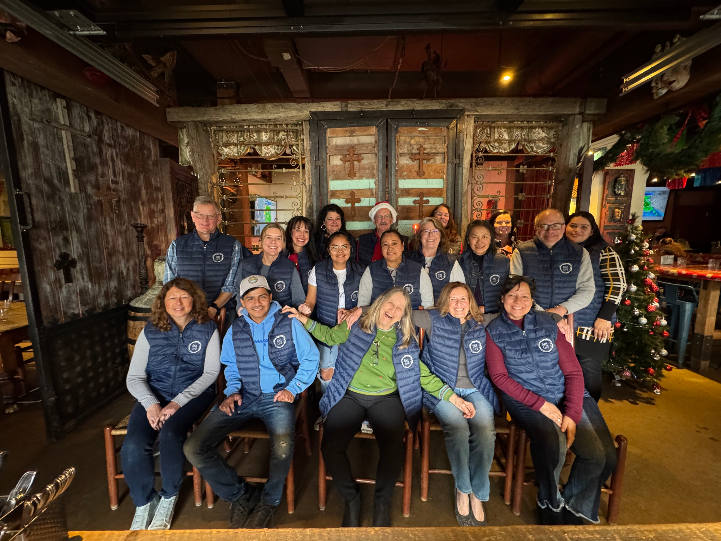 Group of people gathered in a rustic restaurant decorated for Christmas with a Christmas tree, all wearing matching blue jackets.