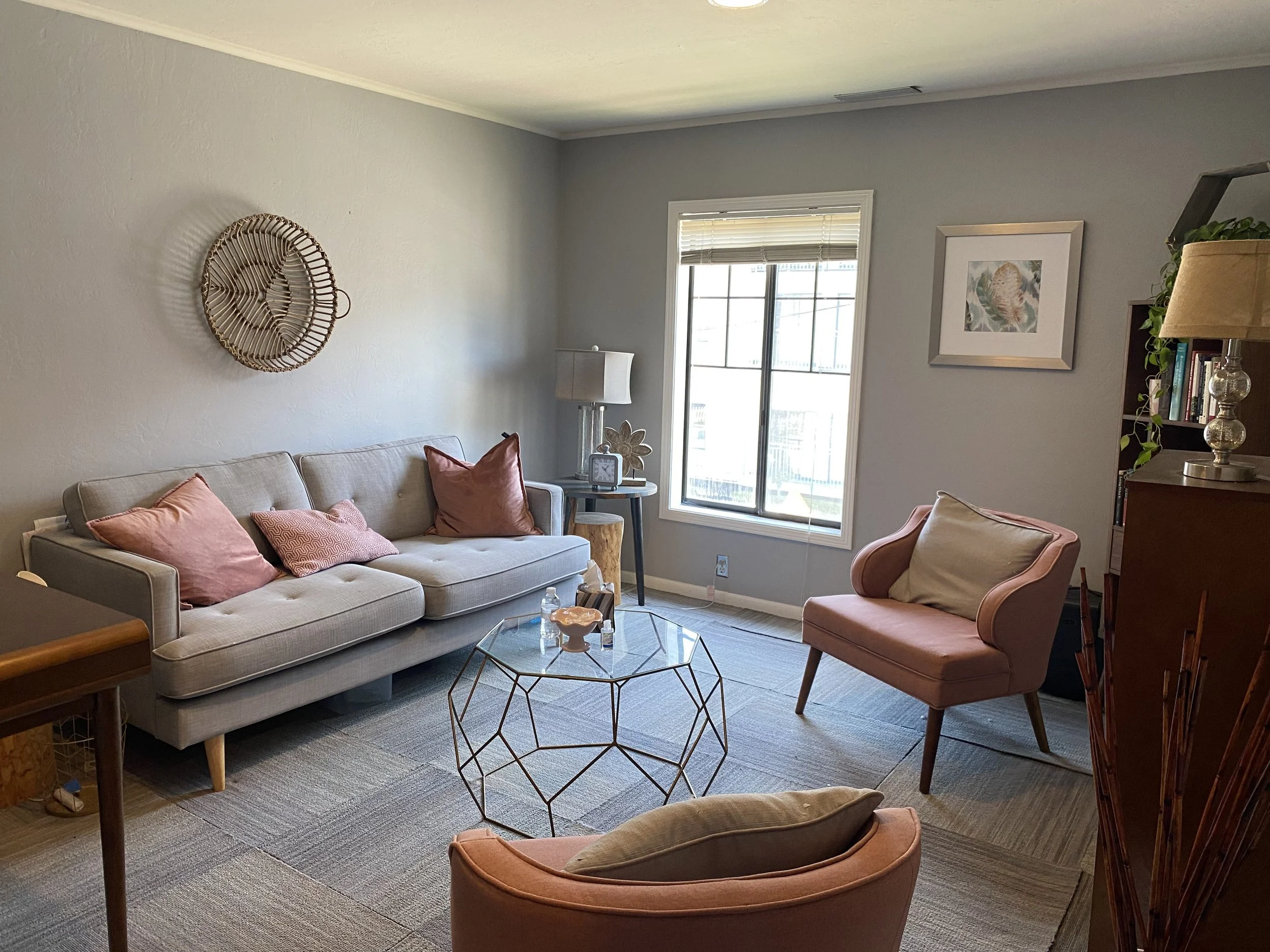 Living room with light gray walls, a beige sofa with pink and purple pillows, a pink armchair, a glass coffee table with a wireframe base, a round end table with a lamp, window with blinds, wall art, and a bookshelf with plants.