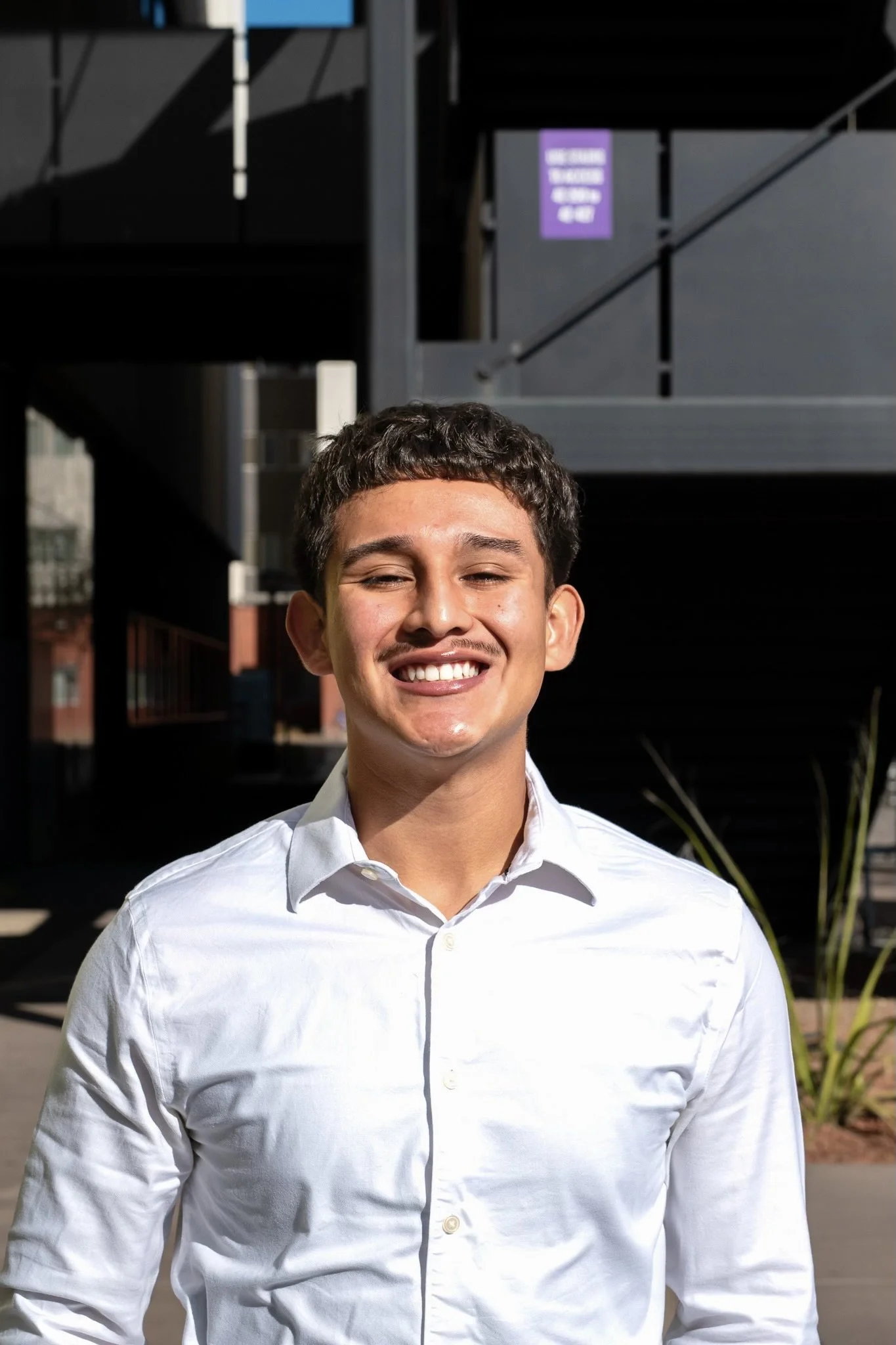 A young man smiling outdoors in front of a modern building, wearing a white collared shirt.