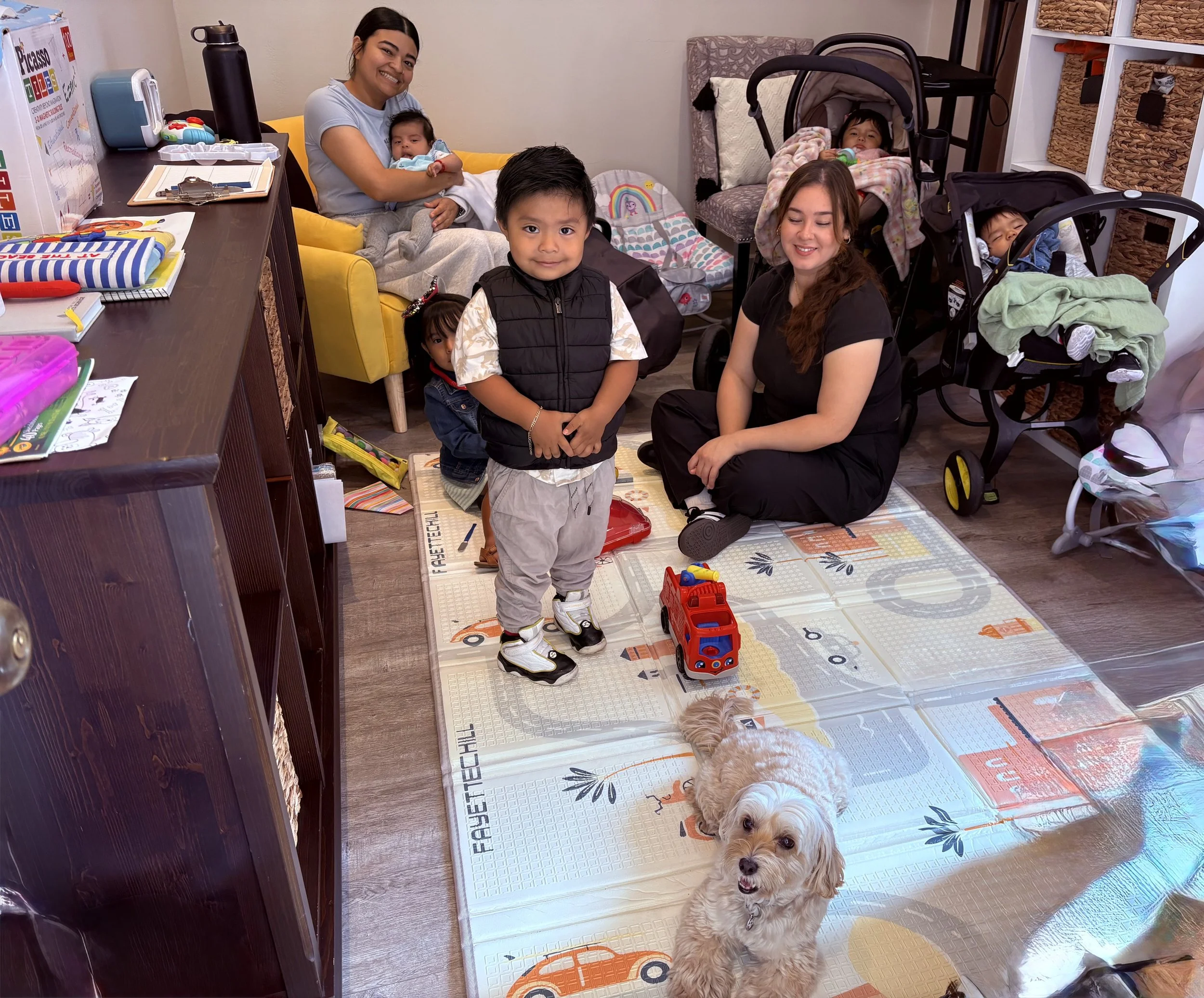 A group of people, including children and adults, gathered in a cozy room with various toys and strollers. A woman is holding a baby, a young boy stands in front of her, and another woman is sitting on the floor smiling. There is a small dog in the foreground on a play mat with colorful designs, and a toy fire truck nearby.