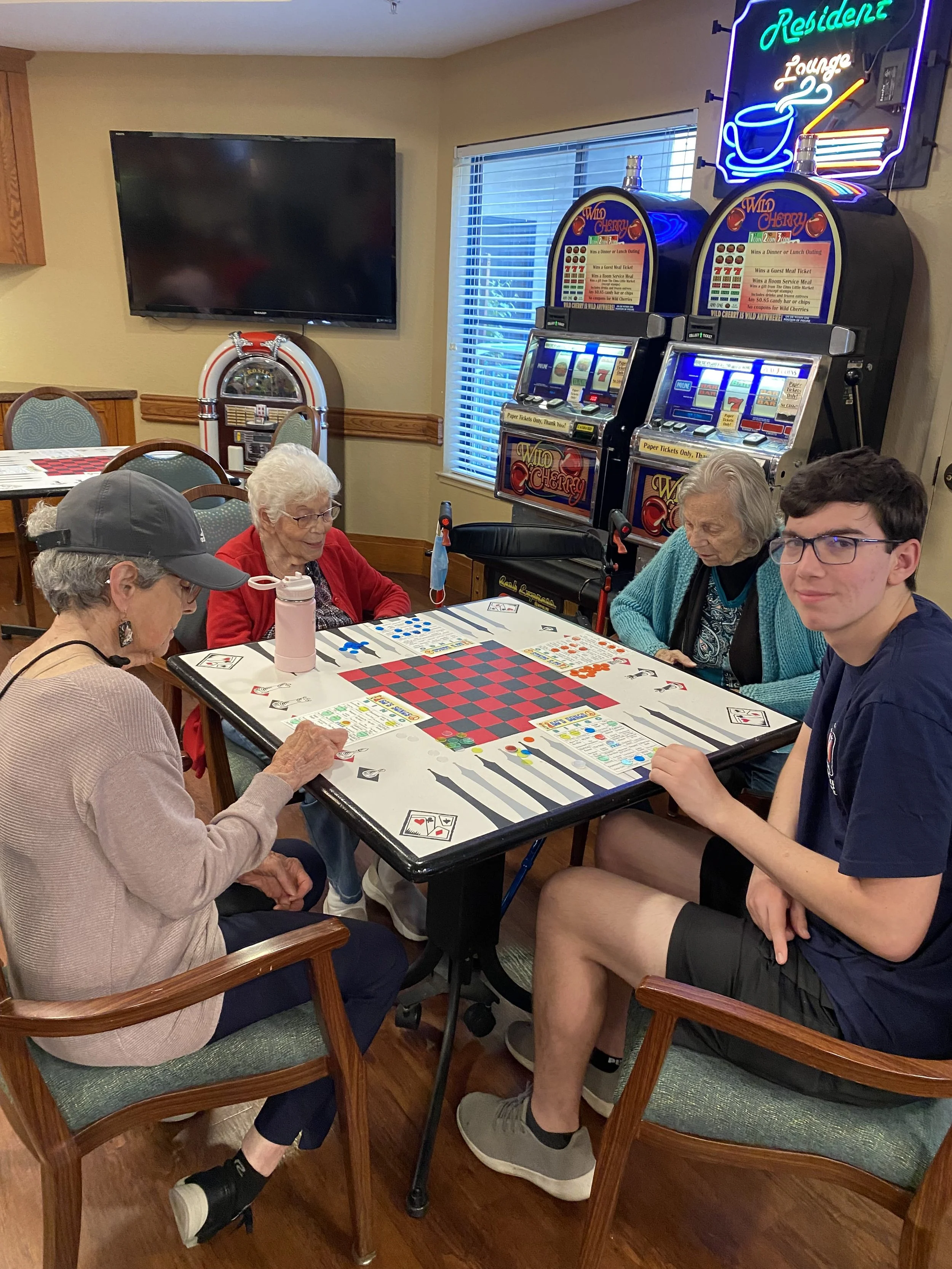 Four people playing checkers at a table in a game room, with slot machines and a neon sign in the background.