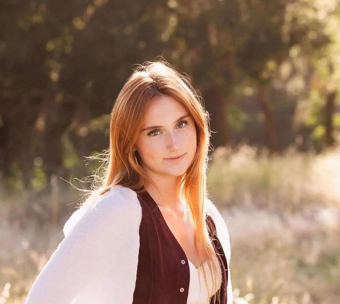 A young woman with long red hair, wearing a white shirt and a black vest, standing outdoors during golden hour with trees and sunlight in the background.