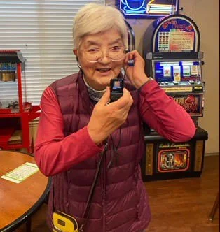 An elderly woman with short white hair talking on a corded phone in a room with a slot machine and a table.