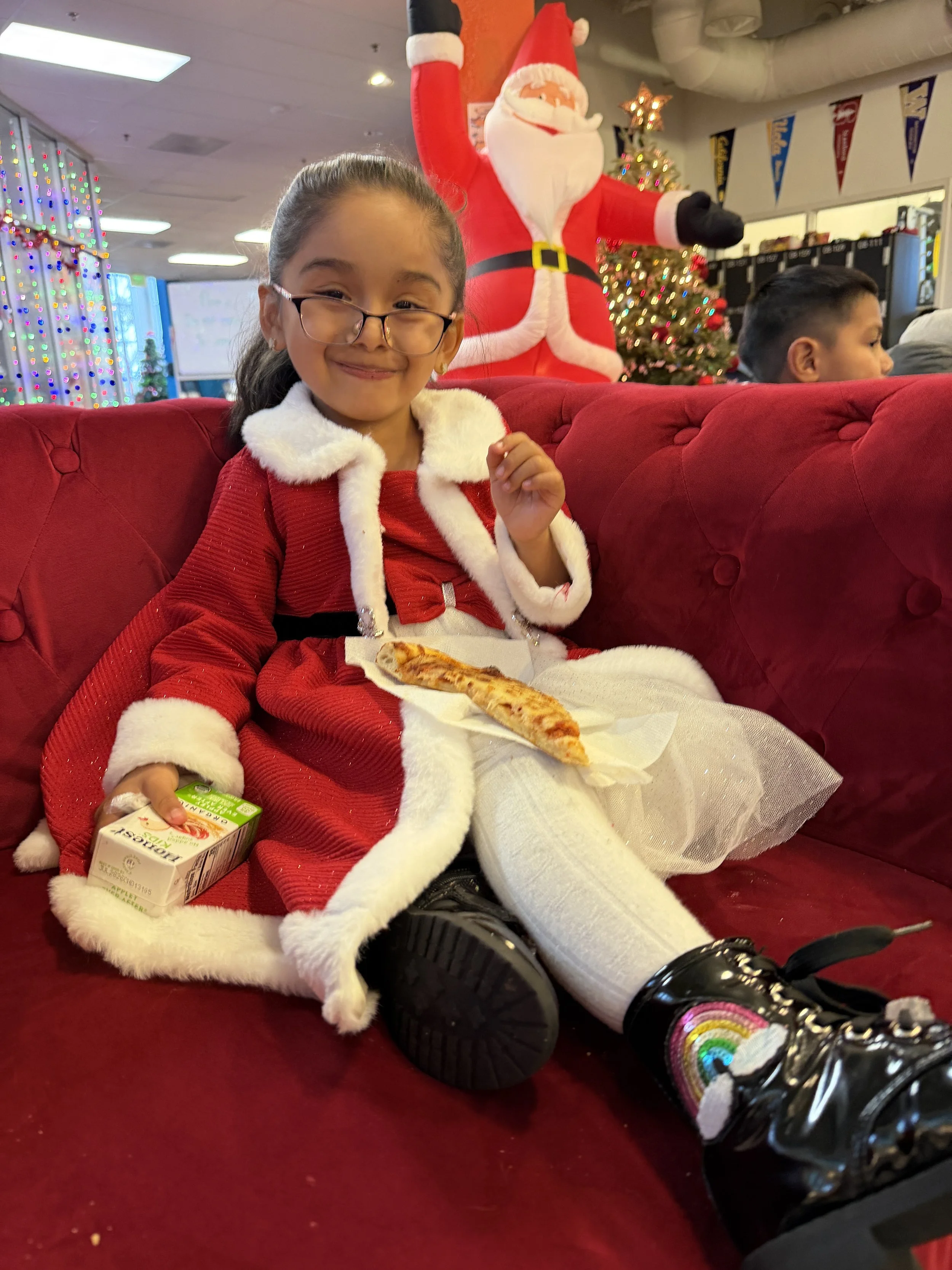 A young girl dressed in a festive Santa outfit sitting on a red velvet couch, holding a juice box and a slice of pizza with Christmas decorations and a large Santa Claus figure in the background.