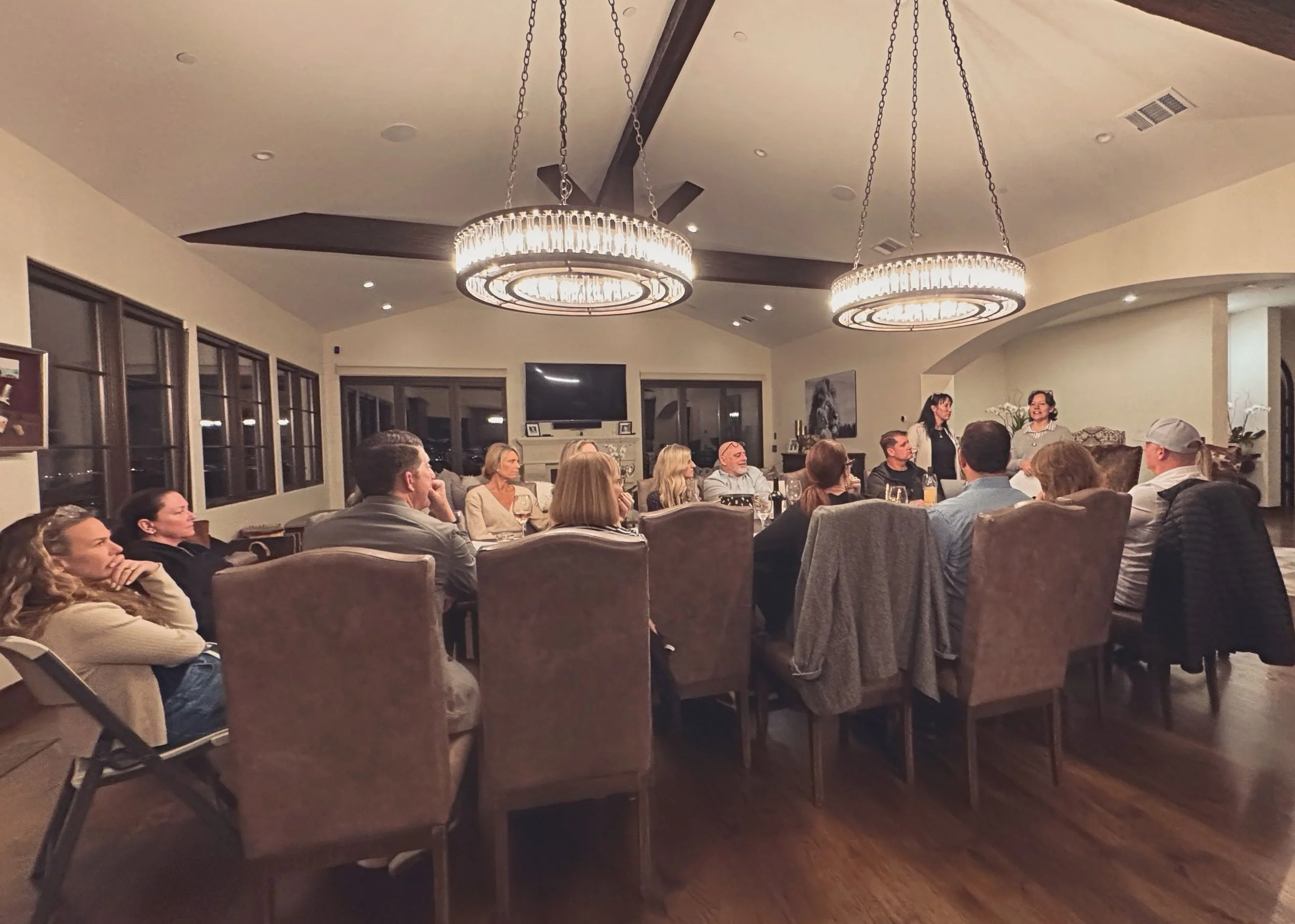 A group of people seated around a large dining table in a well-lit room with large windows, chandeliers, and a television on the wall, attending a presentation or discussion.