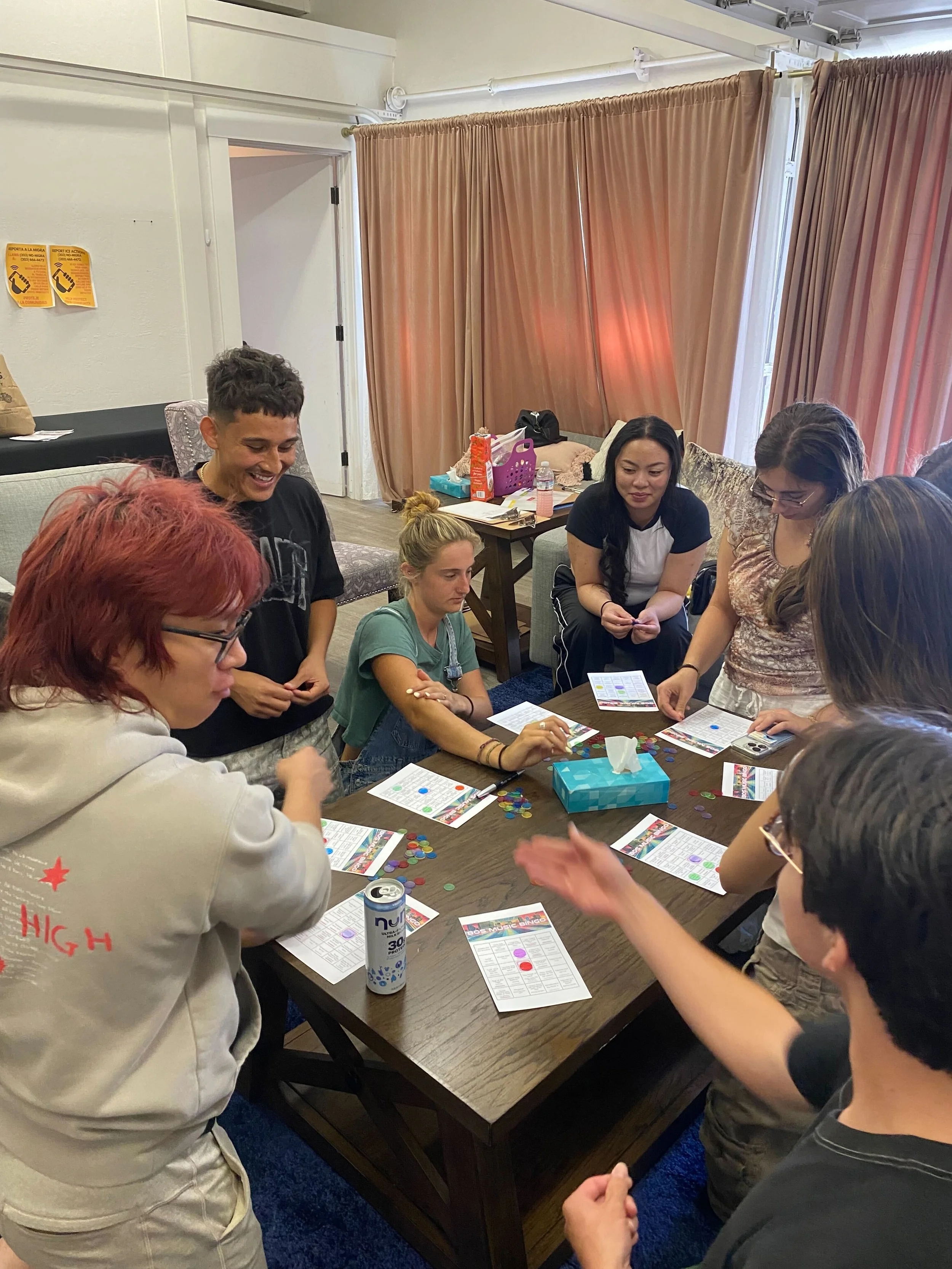 Group of people playing a game around a table in a cozy room with pink curtains.