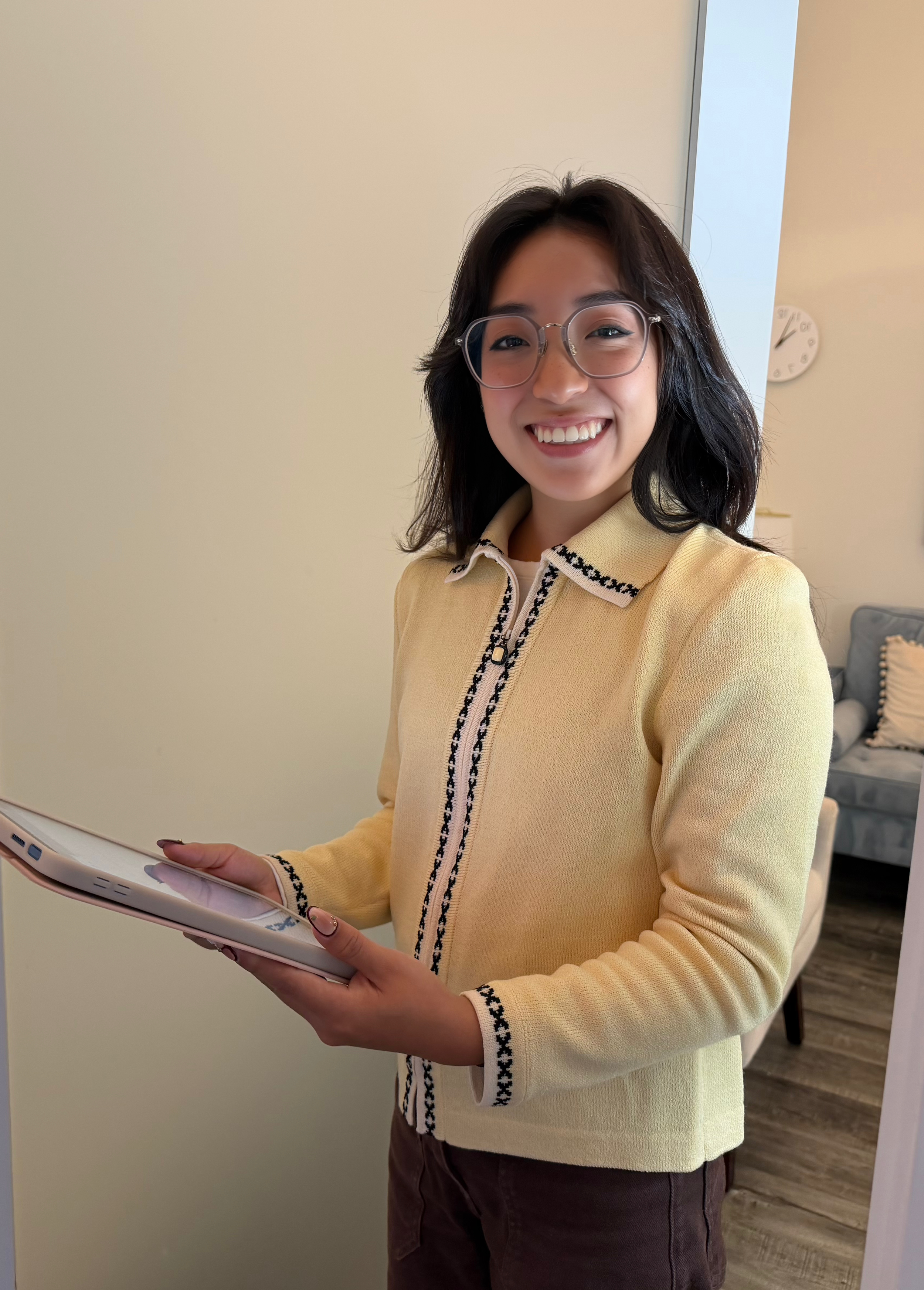 A young woman with shoulder-length dark hair and glasses smiling while holding a tablet in an indoor setting.