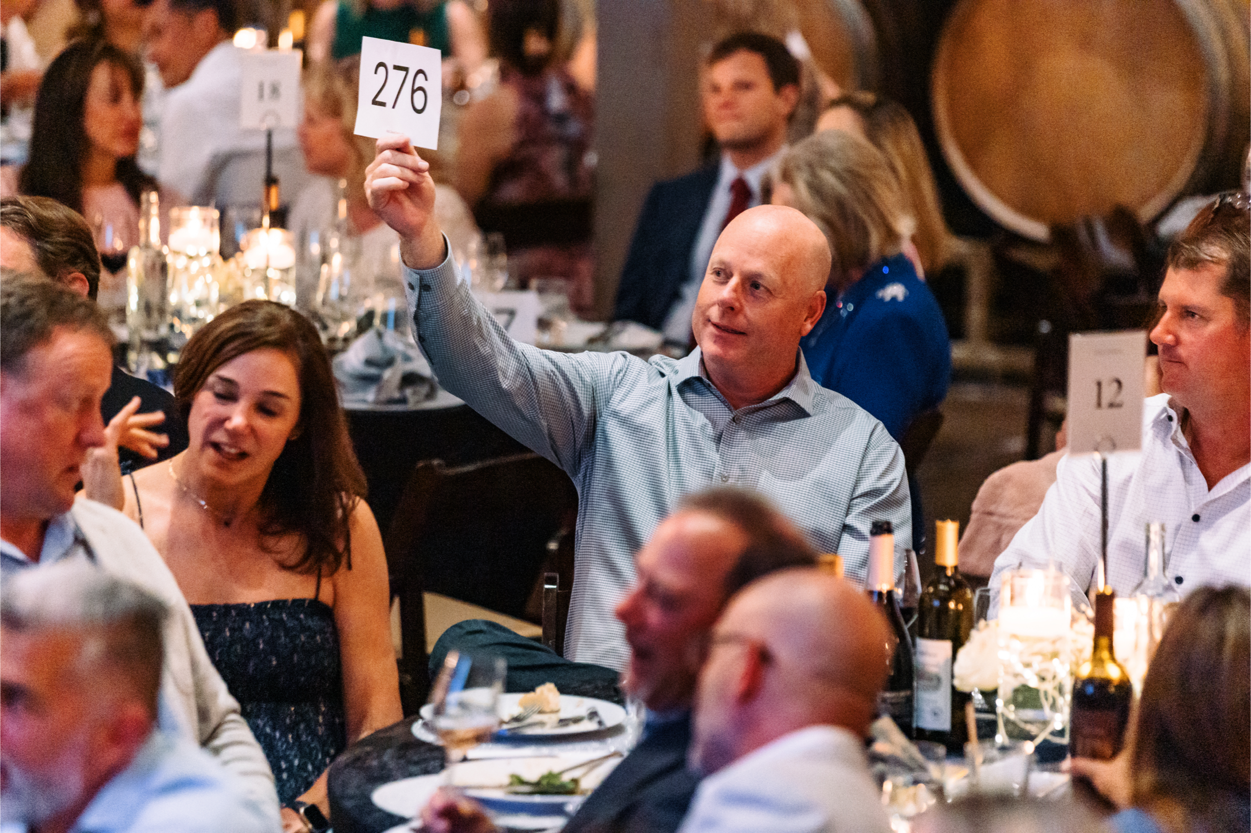 A man with a bald head wearing a light checkered shirt is holding up a bidding paddle with the number 276 at a dinner event, surrounded by other seated attendees at a decorated table with wine bottles and glasses.