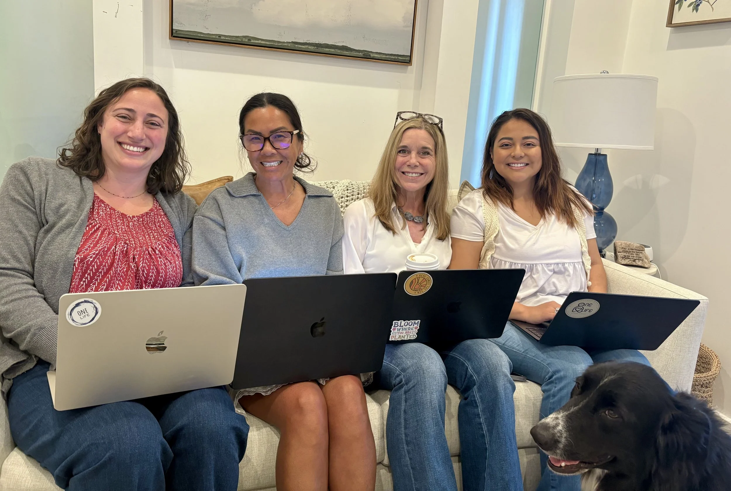 Four women sitting on a sofa with laptops, smiling, with a dog in the foreground and a lamp and artwork in the background.