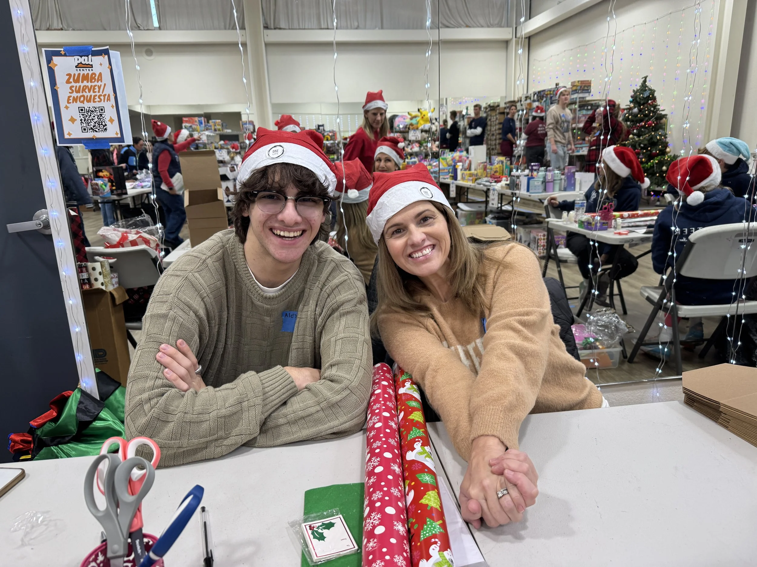Two smiling people in Christmas hats sitting at a table at a holiday event, with gift-wrapping supplies and Christmas presents around them. There are decorated Christmas trees and people working in the background.