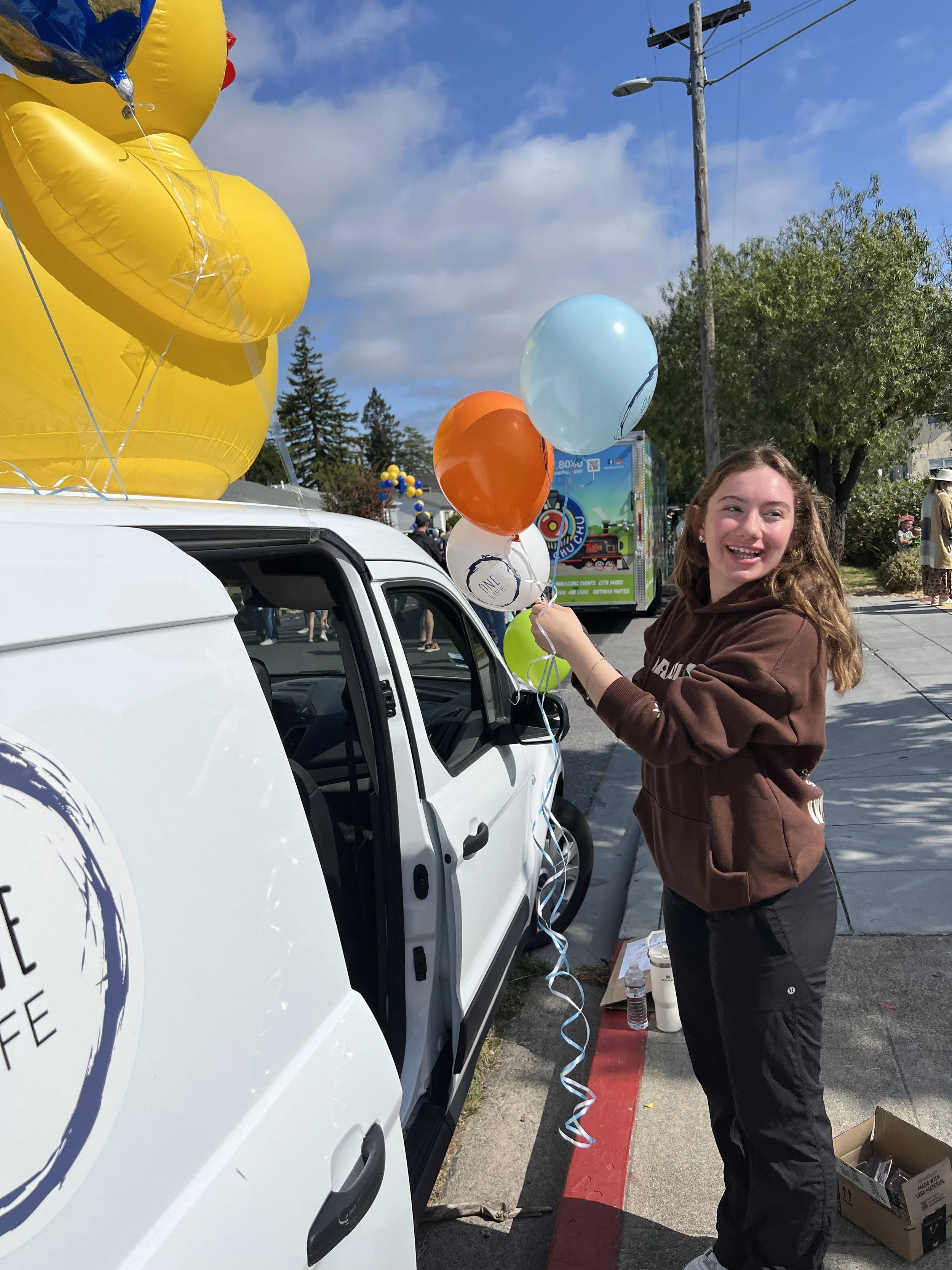 A young girl with long wavy hair is smiling and holding several colorful balloons next to a white vehicle at an outdoor event on a sunny day with a blue sky. There are trees, a utility pole, and other people in the background.