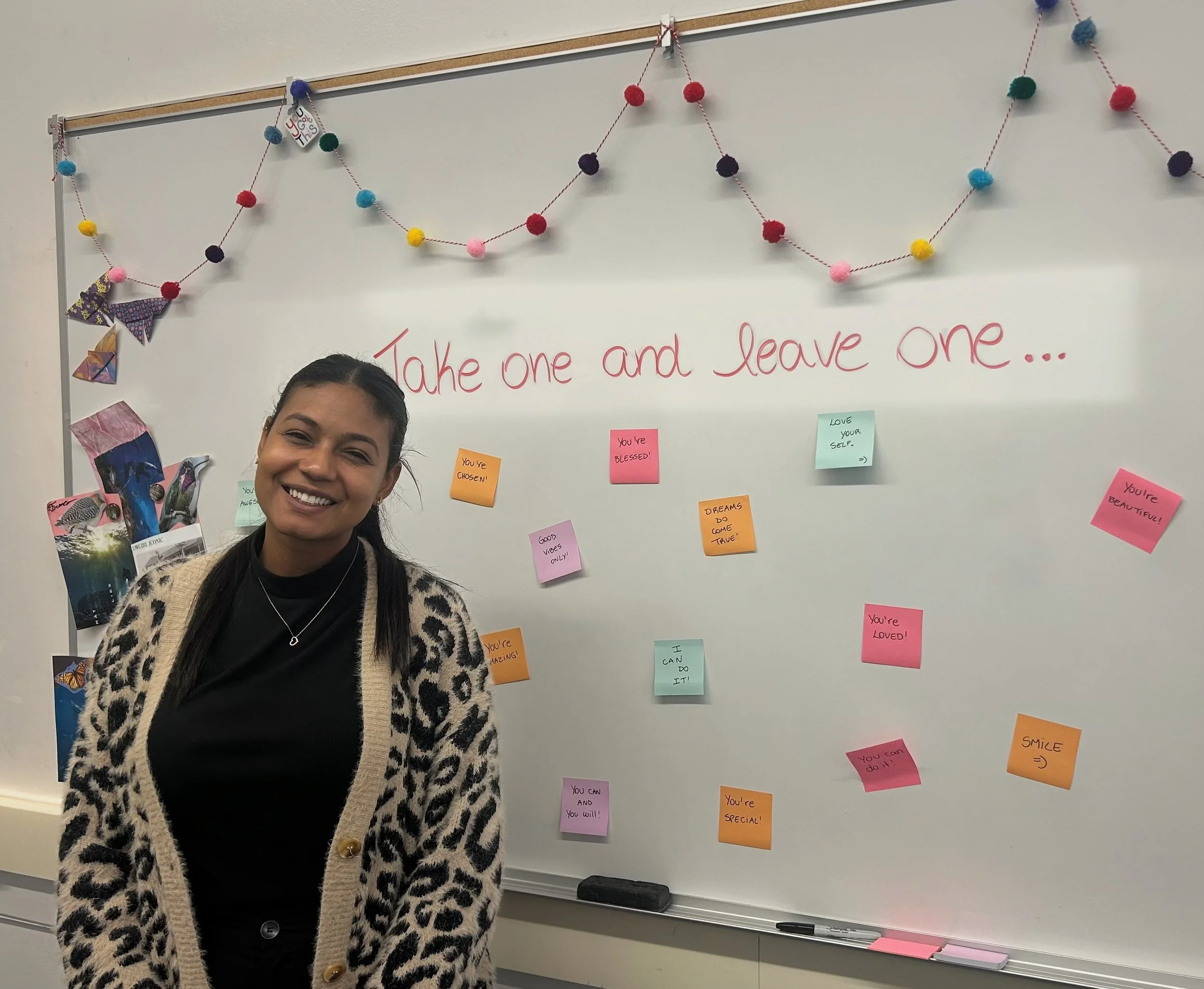 A woman standing in front of a whiteboard with a colorful pom-pom garland and small notes, smiling at the camera.