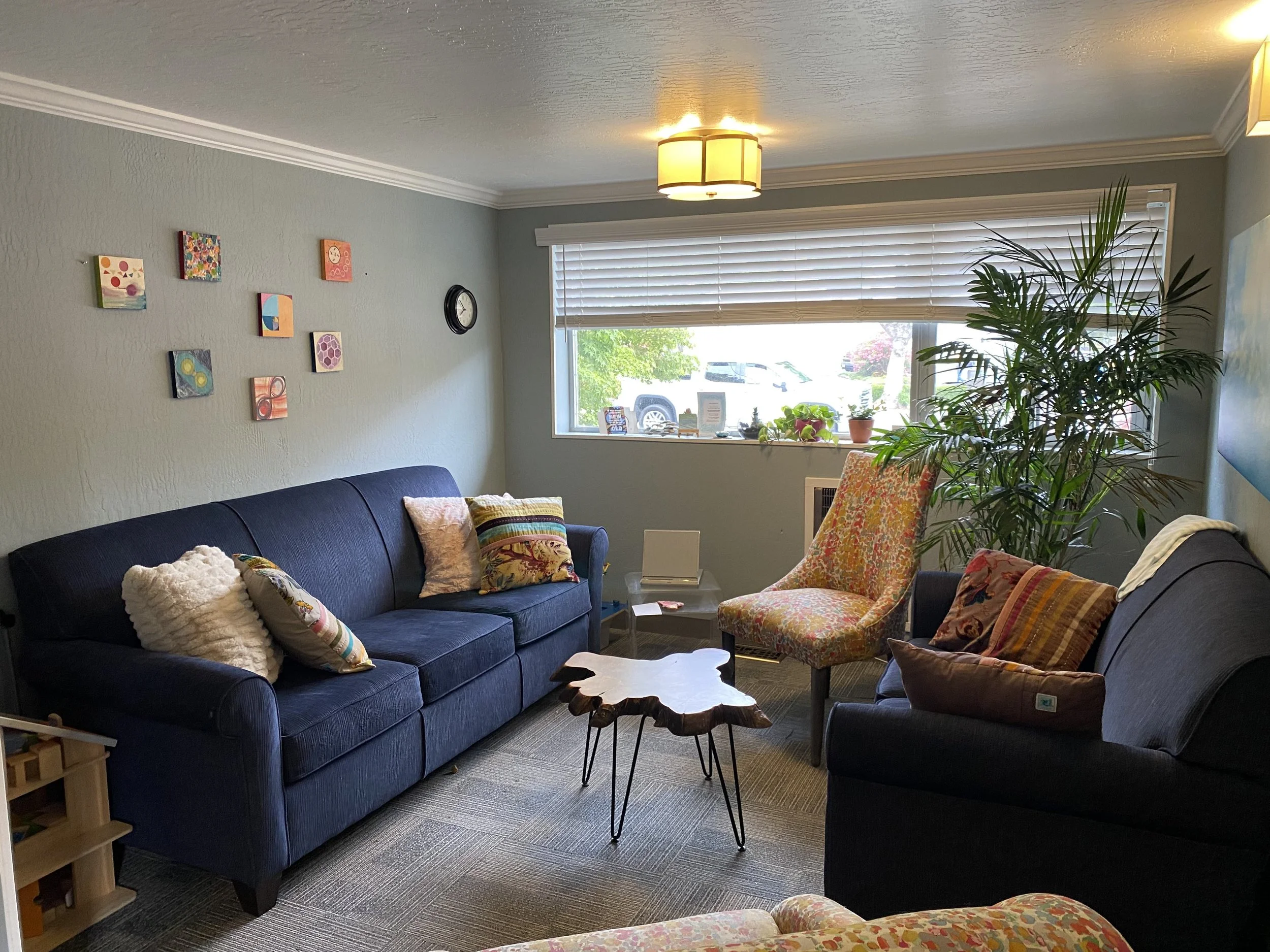 A cozy living room with two navy blue sofas, colorful throw pillows, a small wooden coffee table, a large potted plant, and a window with blinds letting in sunlight. The walls have colorful artwork and a small clock.