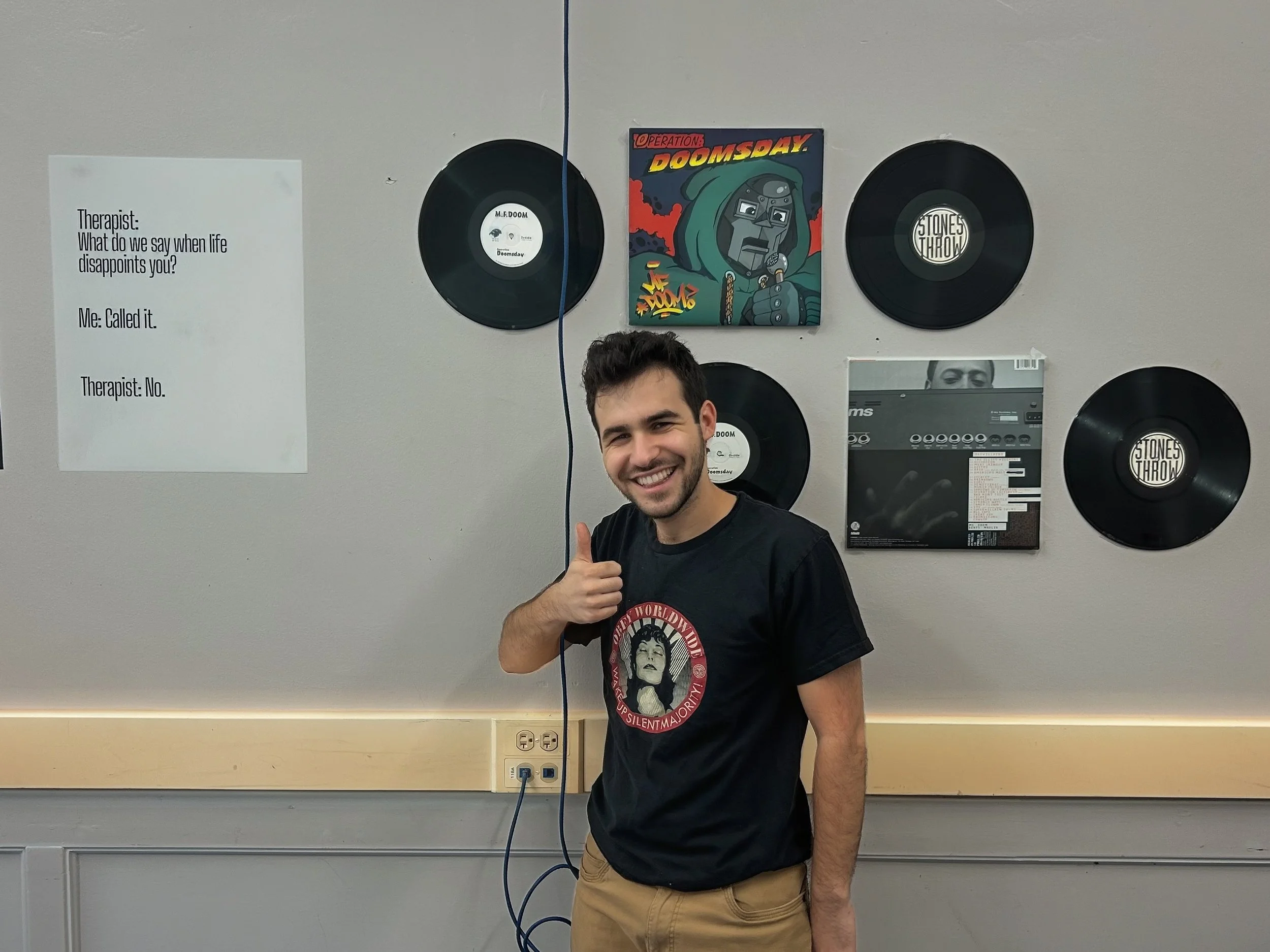A young man with dark hair and a beard, smiling and giving a thumbs-up, standing in front of a decorated wall with vinyl records, album covers, and a framed picture, in what appears to be a music or therapy room.