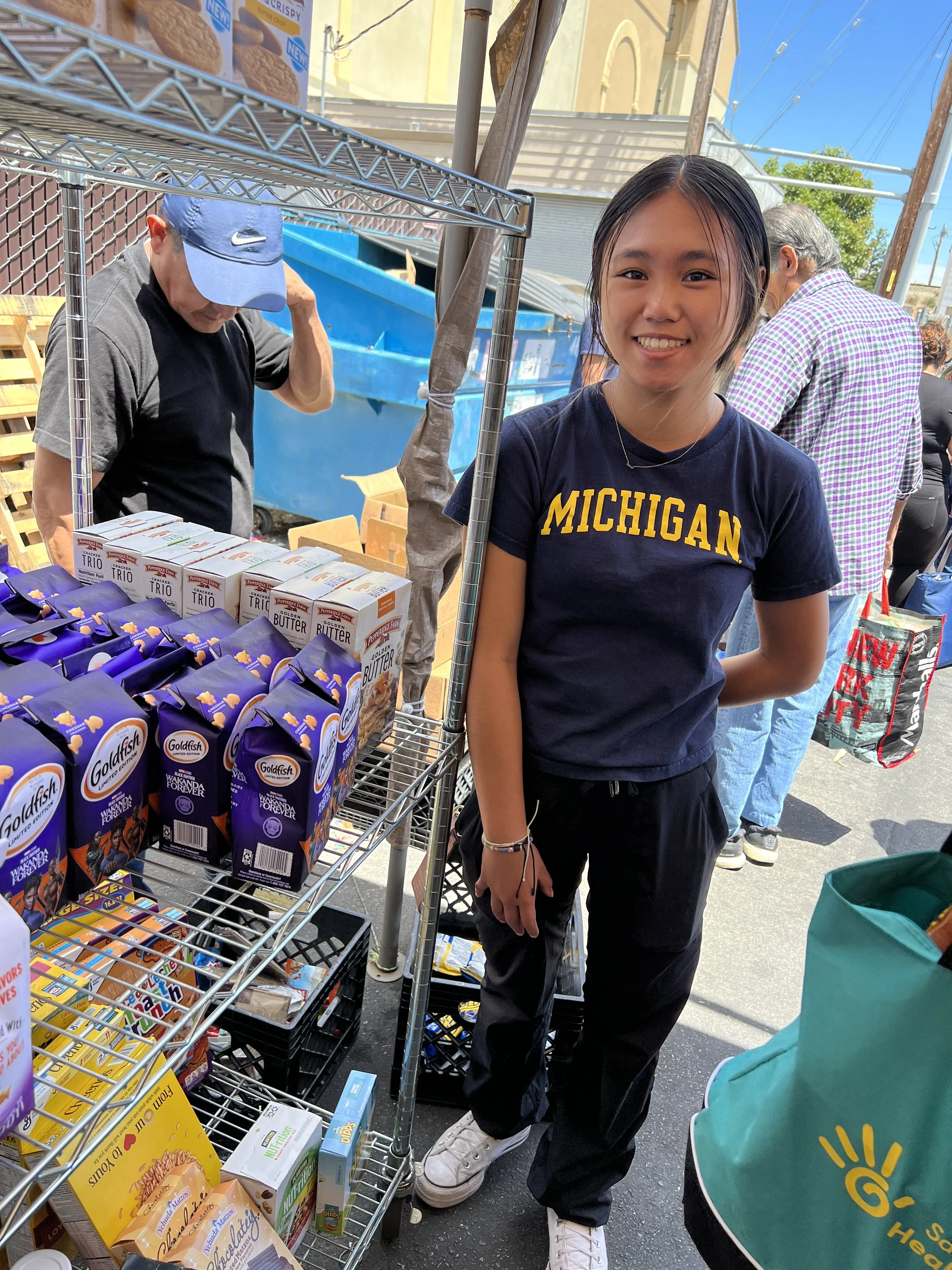 A young woman smiling at the camera, standing next to a shopping cart filled with groceries such as almond milk and cereal at an outdoor market or grocery stand. She is wearing a navy blue Michigan T-shirt, black pants, and white sneakers. There are other people and market stalls in the background.