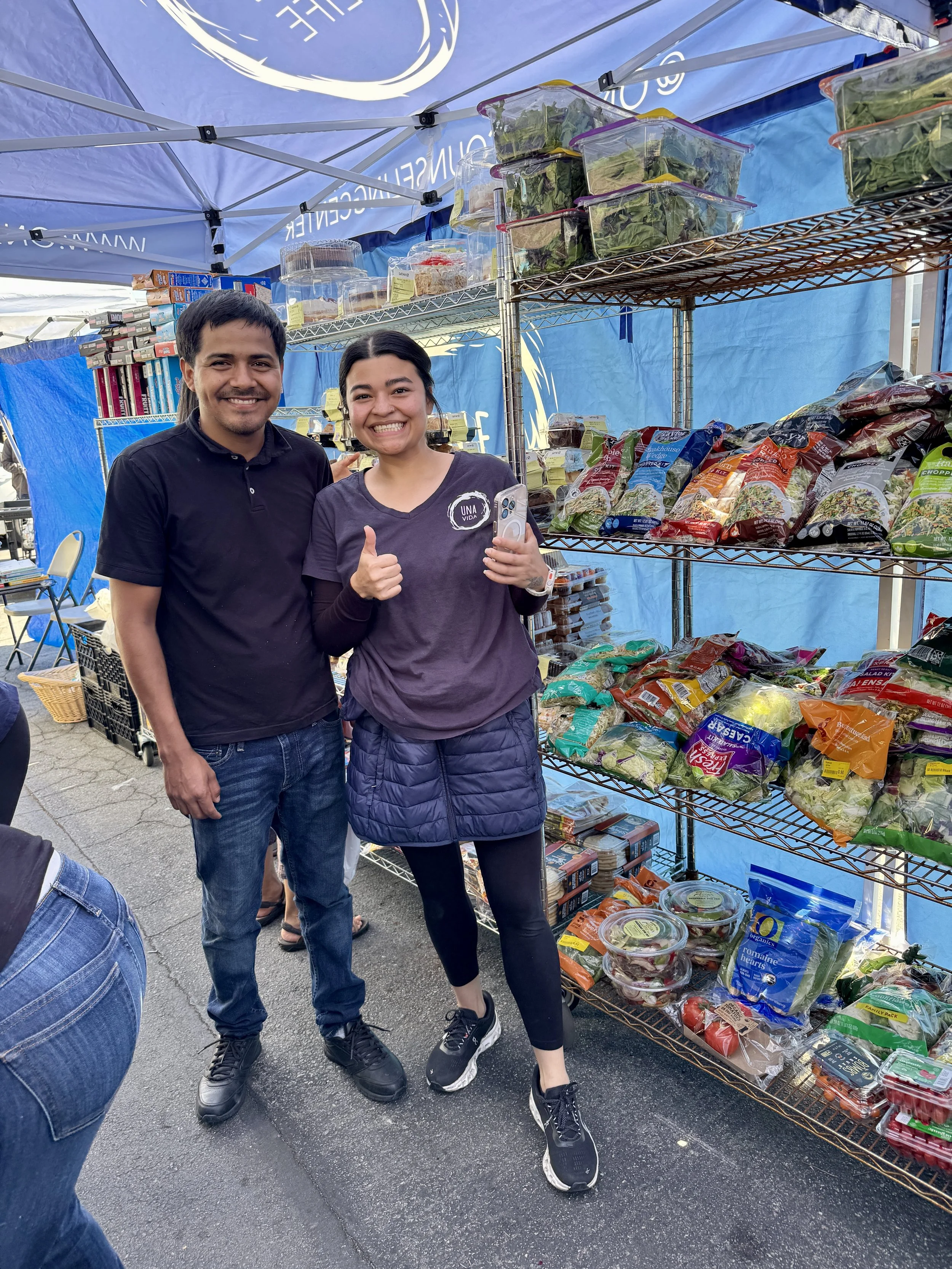 Two people smiling and standing in front of a market stall with shelves stocked with packaged salads and vegetables, outdoors under a blue canopy.