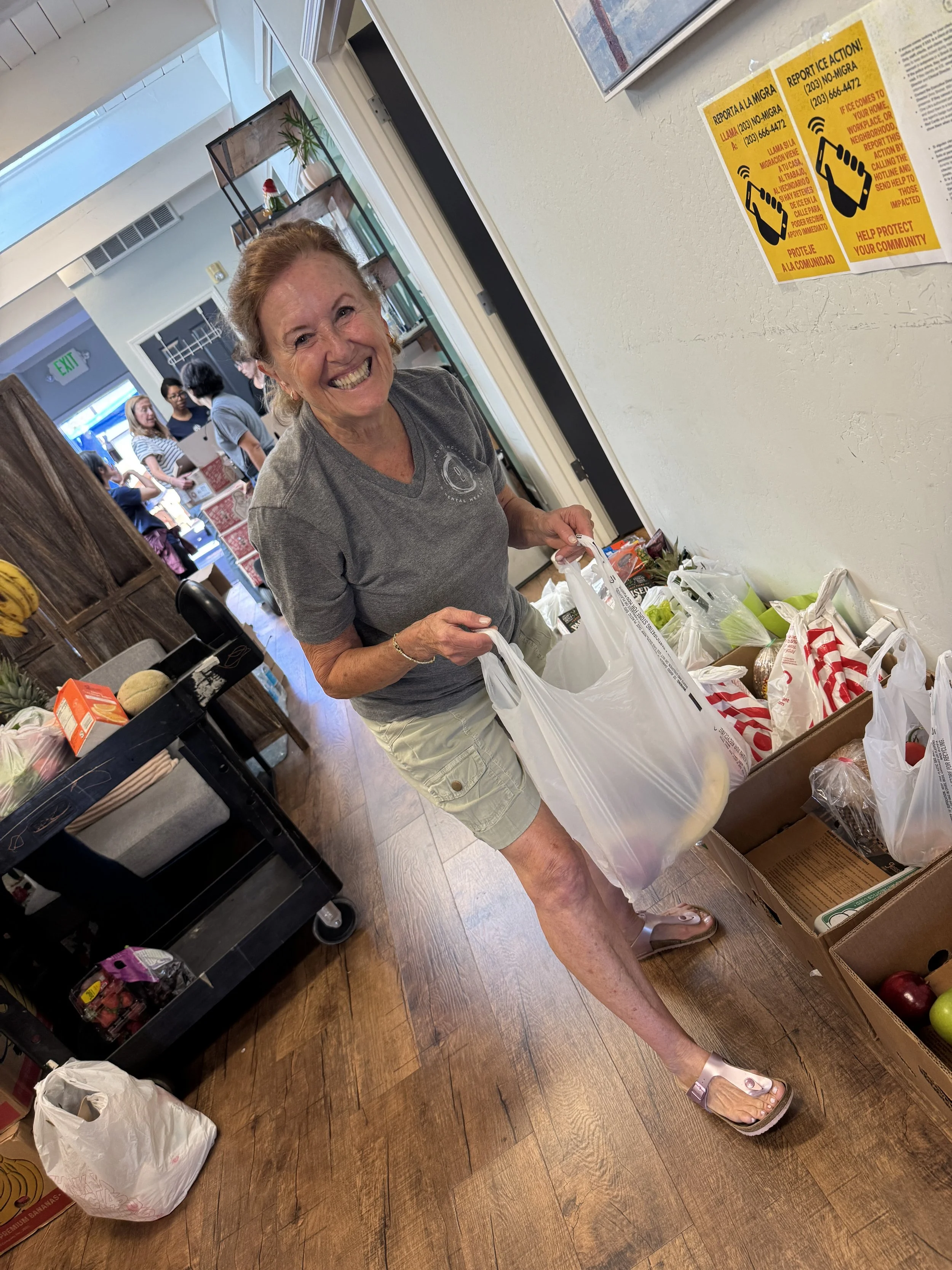 A smiling woman with short hair wearing a gray t-shirt and khaki shorts shopping at a grocery store, holding plastic bags filled with produce and supplies. She is standing near a display of fruits and vegetables on the floor, with other shoppers in the background.