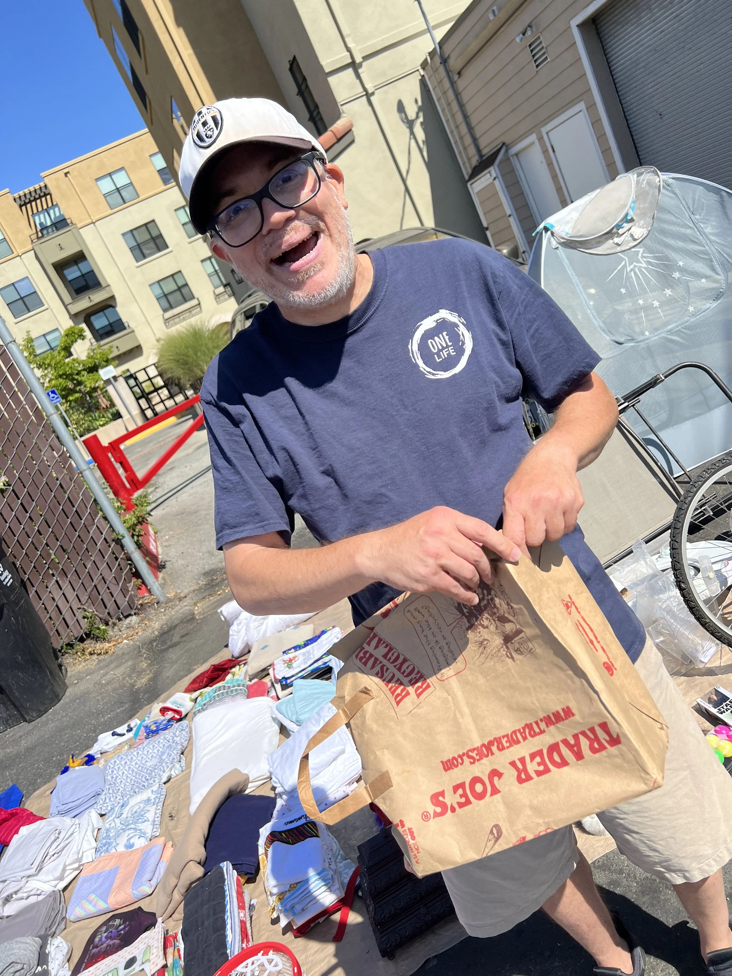 A man with glasses, wearing a beige cap and a navy t-shirt, is smiling and holding a brown paper bag at an outdoor yard sale with various clothes and items on tables around him.