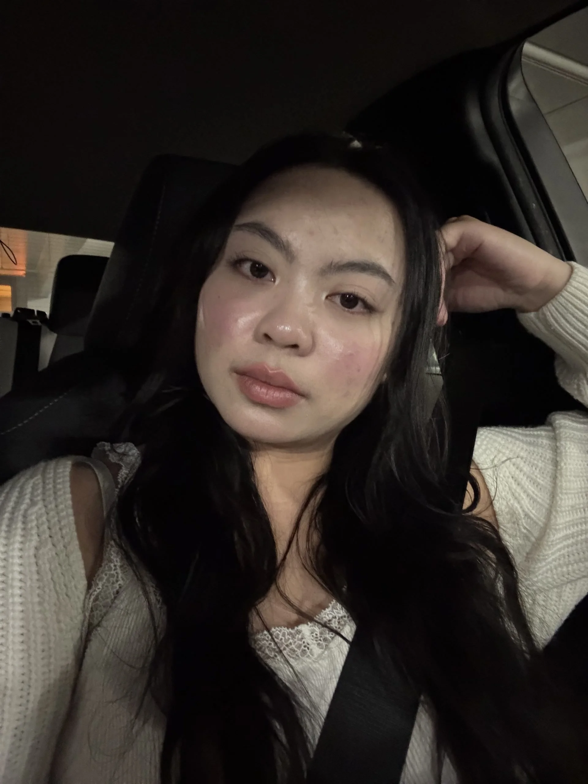 A young woman with long dark hair and light makeup sitting in a car seat, wearing a beige sweater, with her arm resting on her head and looking at the camera.
