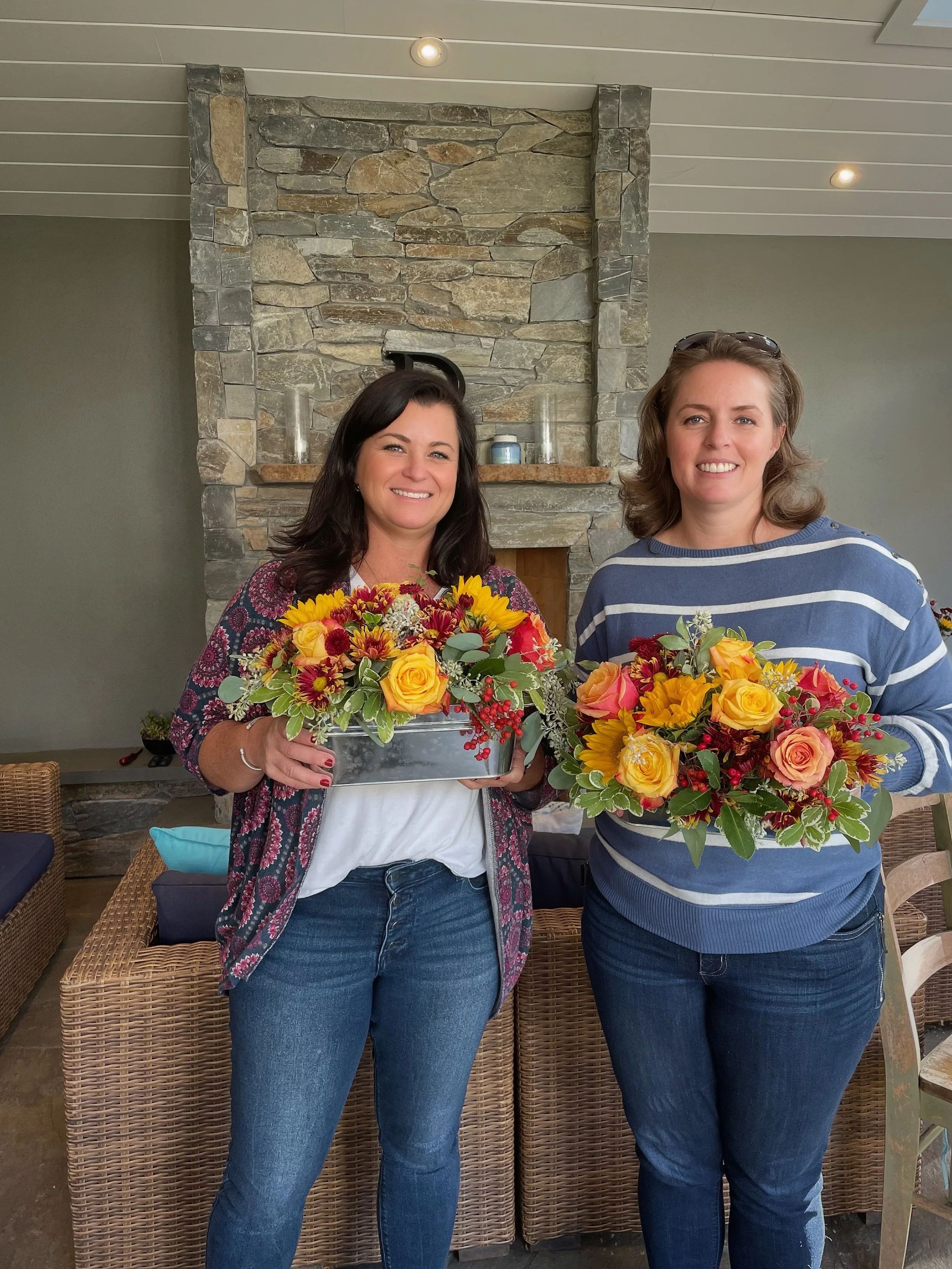Two women standing indoors in front of a stone fireplace, holding colorful bouquets of flowers, smiling at the camera.