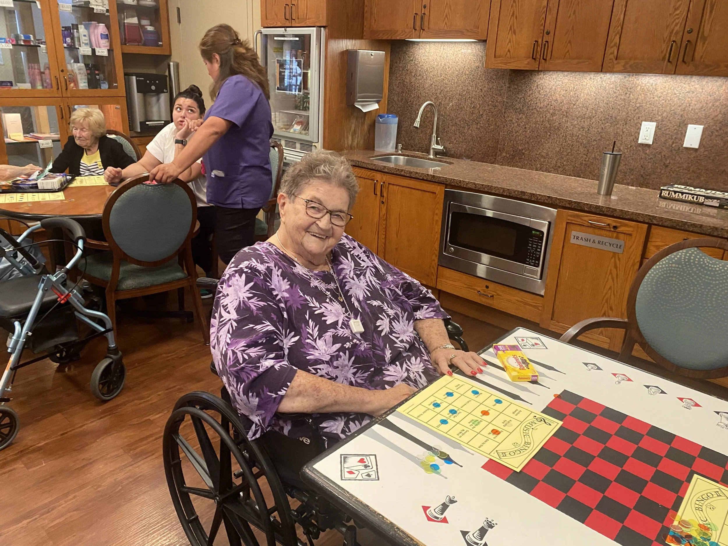 An elderly woman in a wheelchair playing checkers at a table with a bingo card, game pieces, and candy, smiling at the camera. In the background, there are other people, including a caregiver and a woman with a walker, in a room with wooden cabinetry