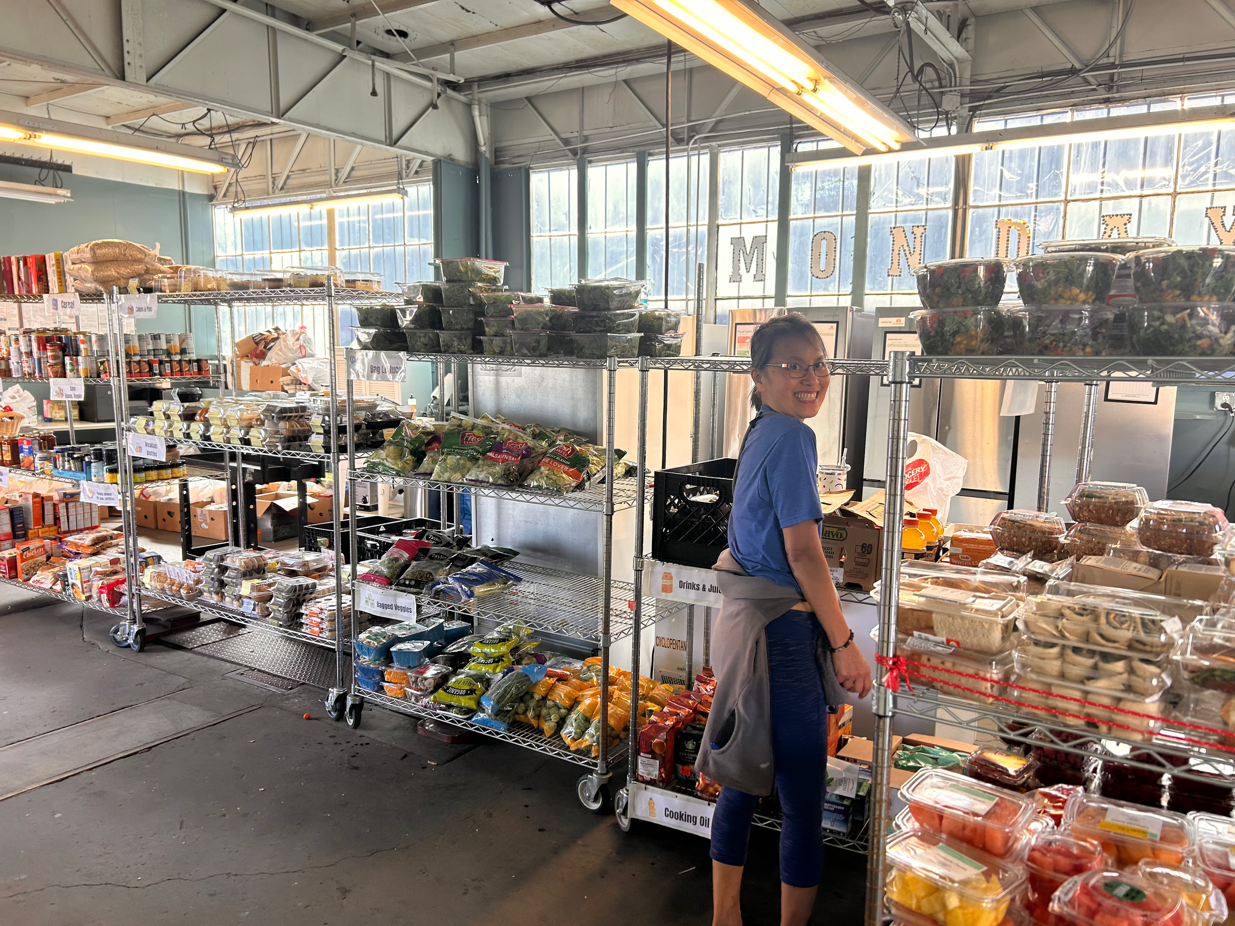 A woman shopping at a grocery store with metal shelves filled with pre-packaged salads, vegetables, and other food items, smiling at the camera, in an industrial-looking store with large windows and natural light.