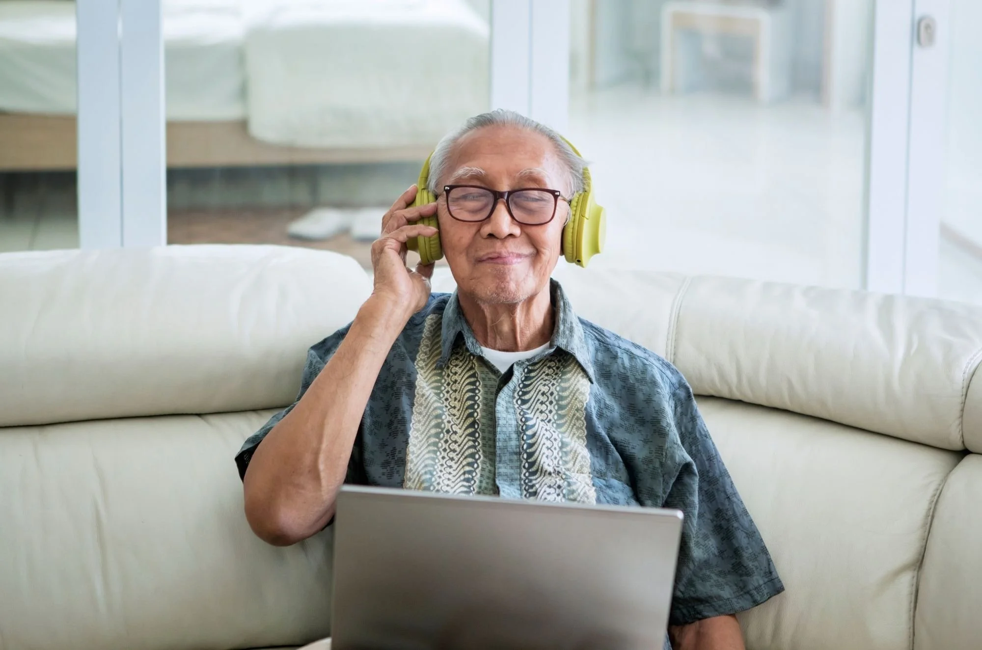 An elderly man sitting on a white sofa, wearing glasses and headphones, listening to music or a podcast with a content smile, with a laptop in front of him.