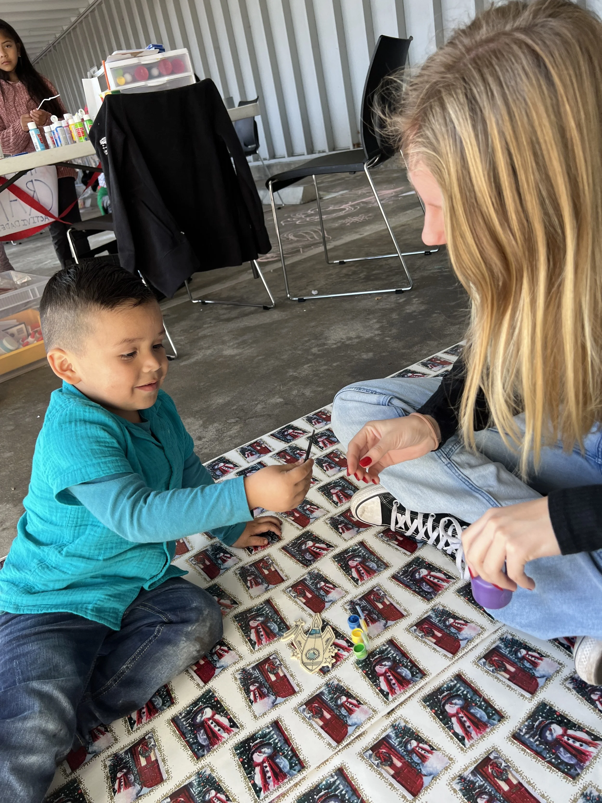 A young boy and a woman sitting on a festive tablecloth decorated with Christmas cards, playing a board game with colorful game pieces.