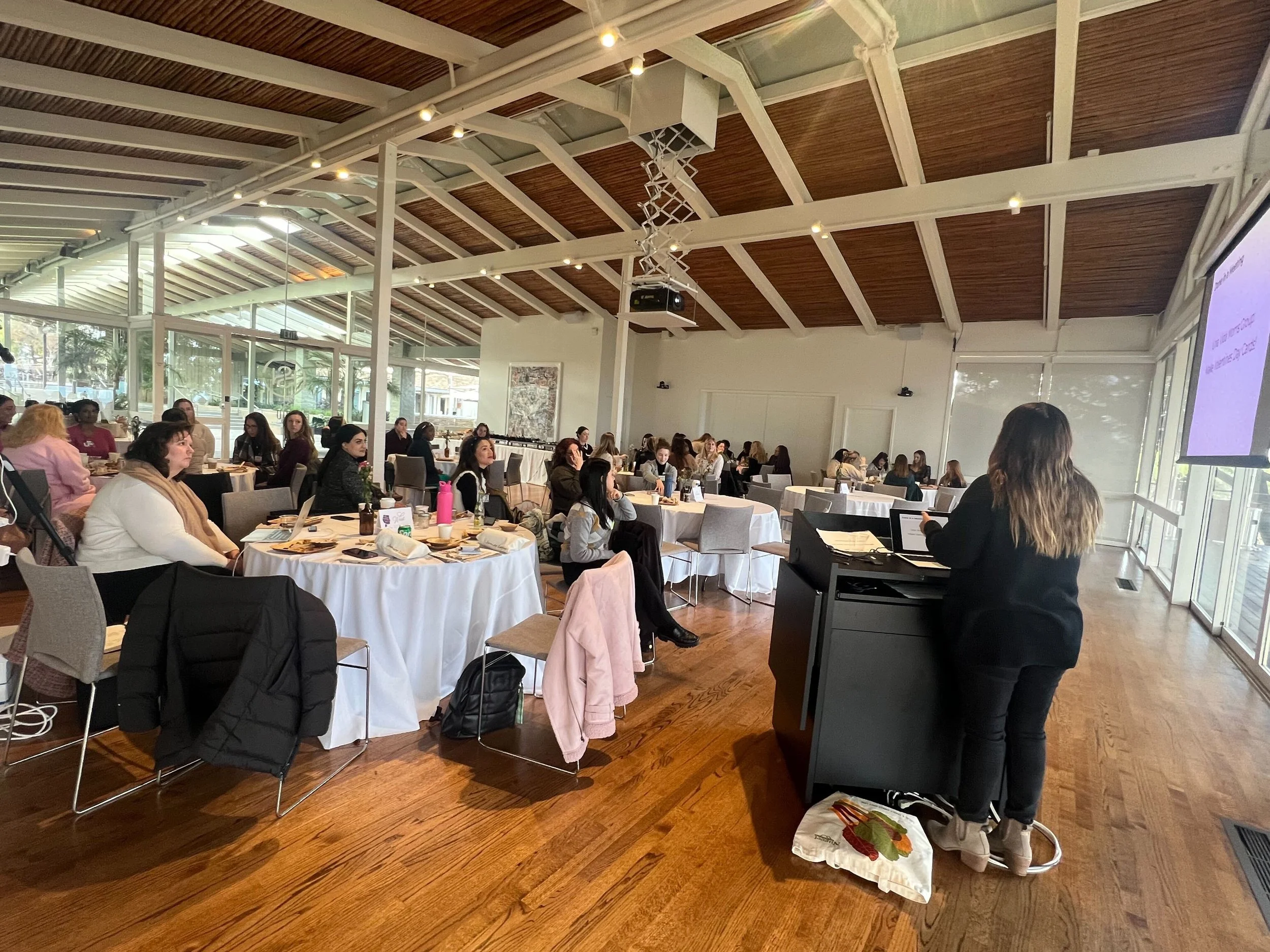A woman giving a presentation to a room full of seated attendees at round tables, in a well-lit space with large windows and a wooden ceiling.