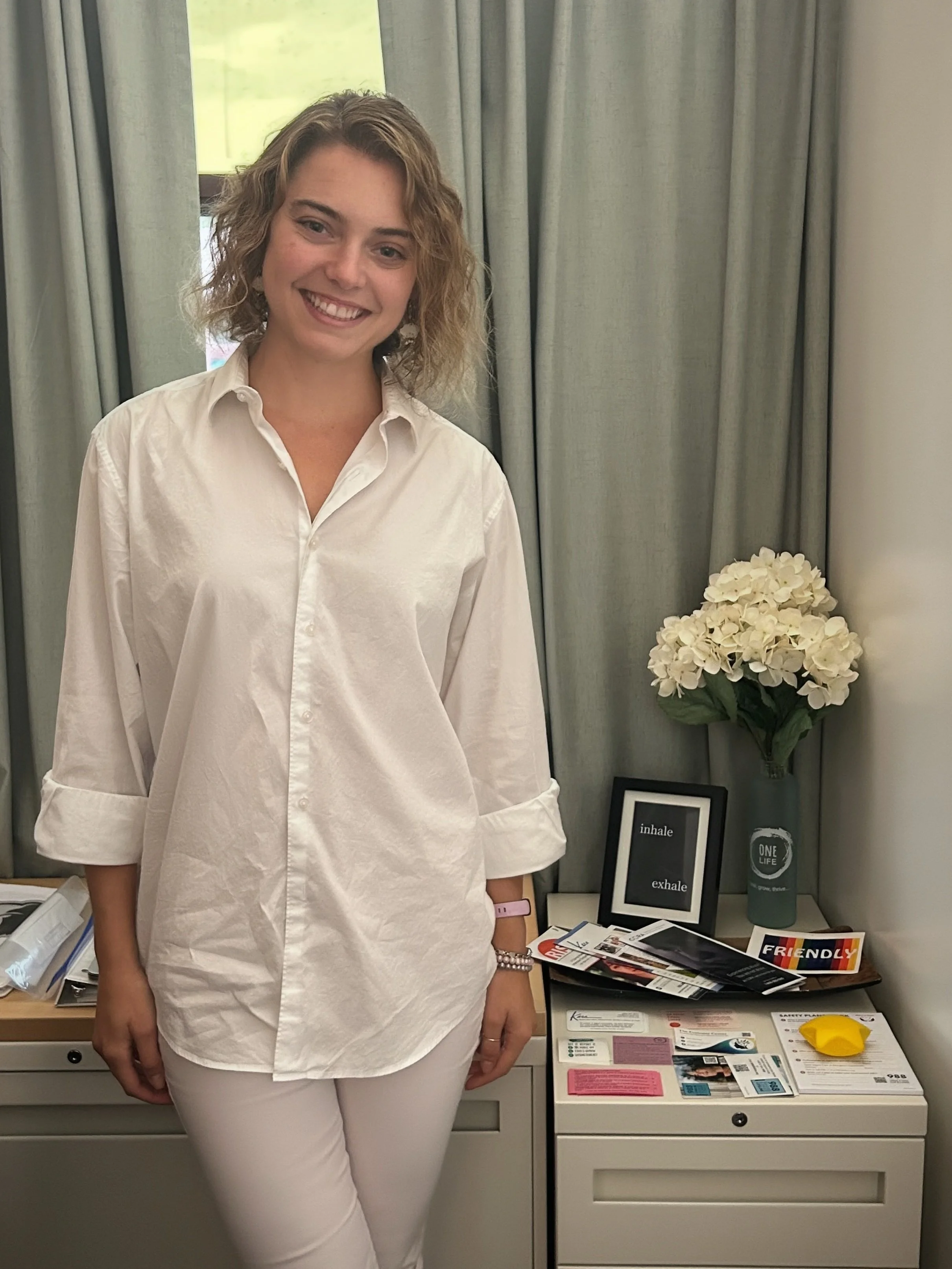 A woman with curly blonde hair smiling, standing in front of a window with greenish curtains, wearing a white shirt and white pants, with a pink wristband and beaded bracelet, standing beside a table with a vase of white flowers and various pamphlets