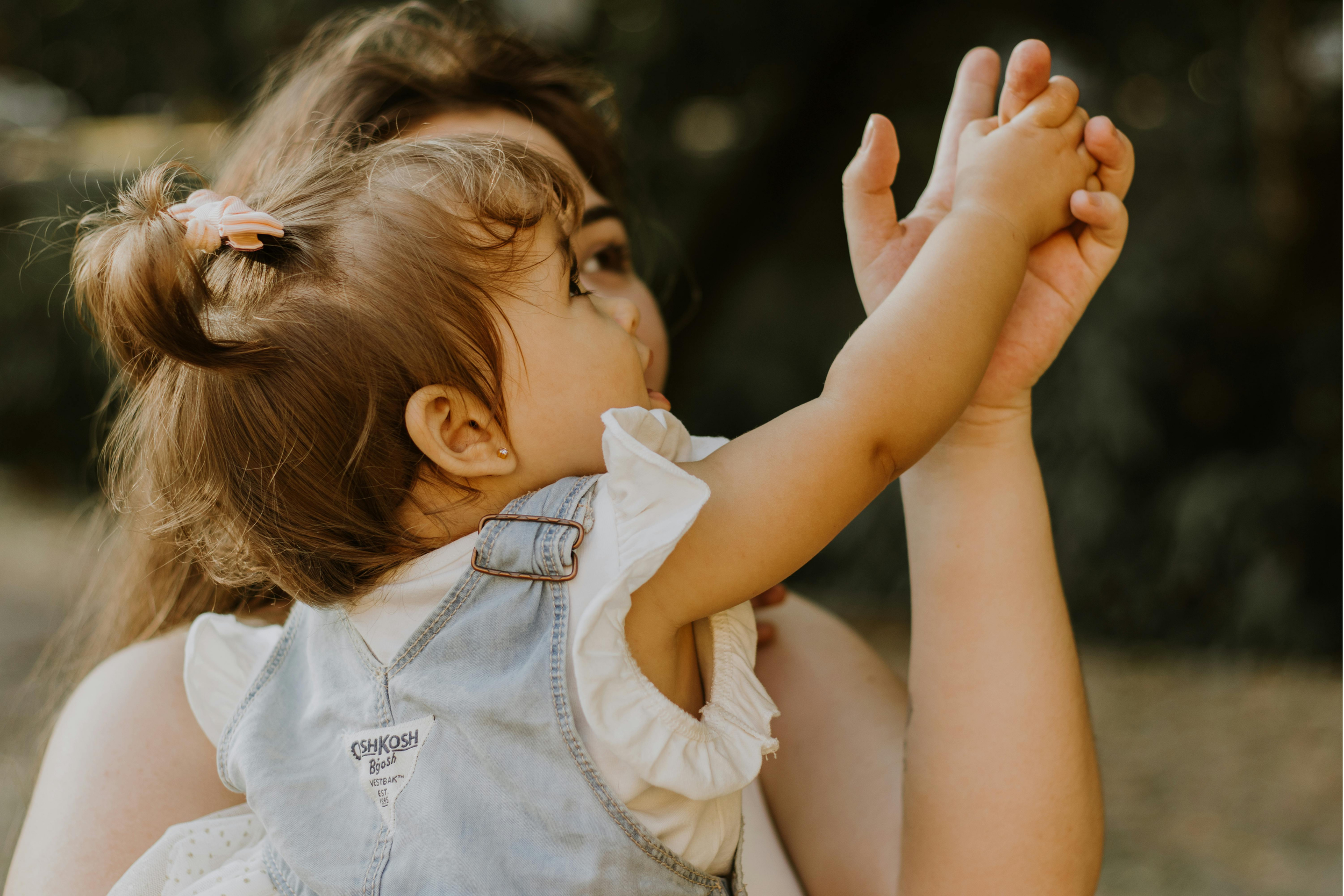 A young girl and woman, possibly her mother, are holding hands outdoors. The girl has brown hair tied with a pink clip, and they are both looking at their clasped hands with a blurred natural background.
