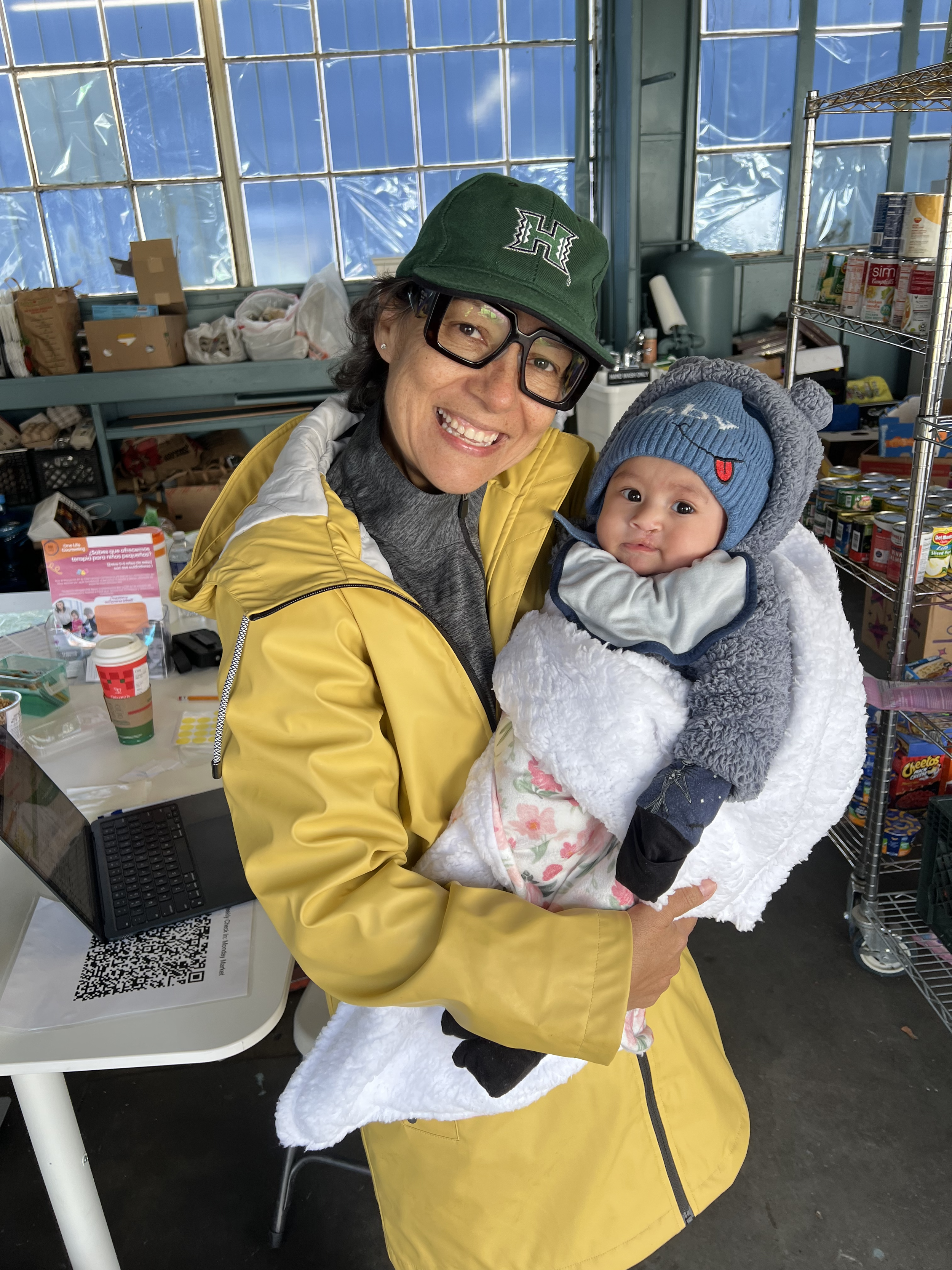 Smiling woman in a yellow raincoat holding a baby dressed in warm clothes, inside a room with shelves and supplies.