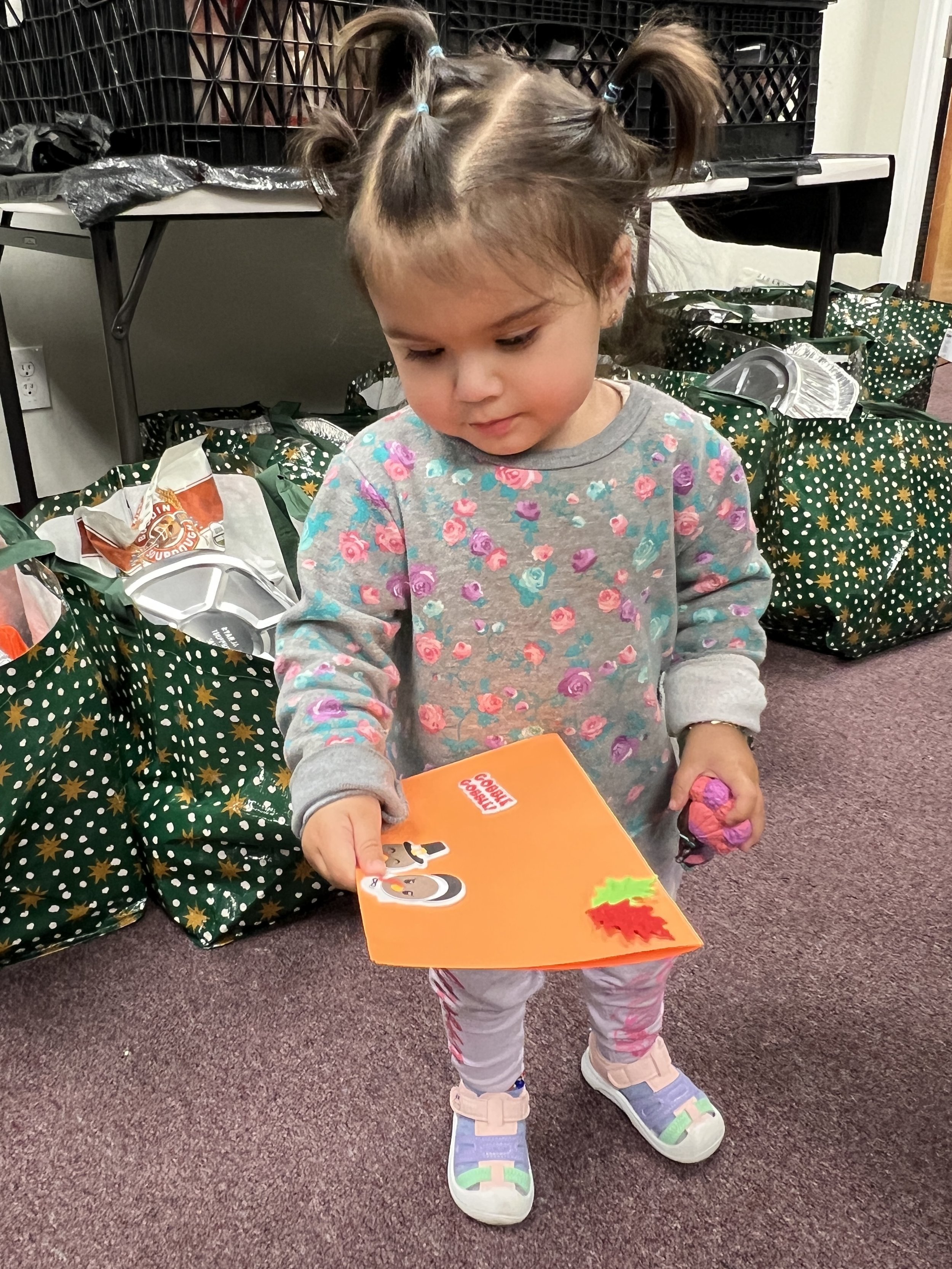 A young girl, with her hair styled in two pigtails, is holding a book with a cartoon dog on the cover, standing among groups of gift bags filled with presents on a carpeted floor.