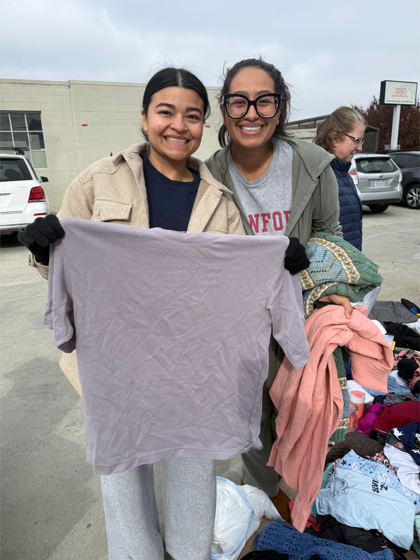 Two women smiling and holding up a wrinkled light purple T-shirt at a thrift or swap meet in a parking lot, with clothes and other items on tables nearby.