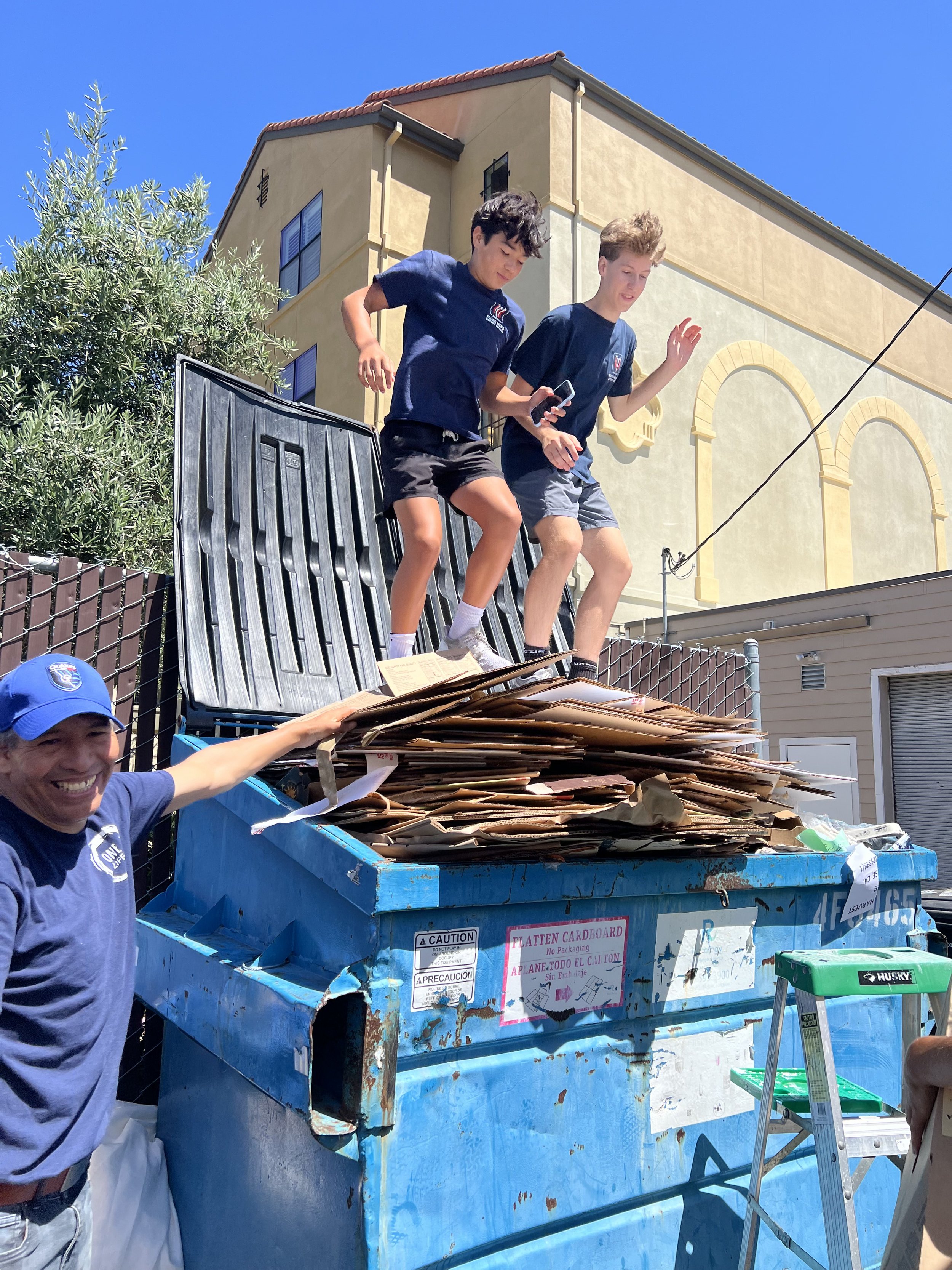 Two young boys jumping off a dumpster filled with cardboard boxes while a man smiles nearby. The scene is outdoors with a building and blue sky in the background.