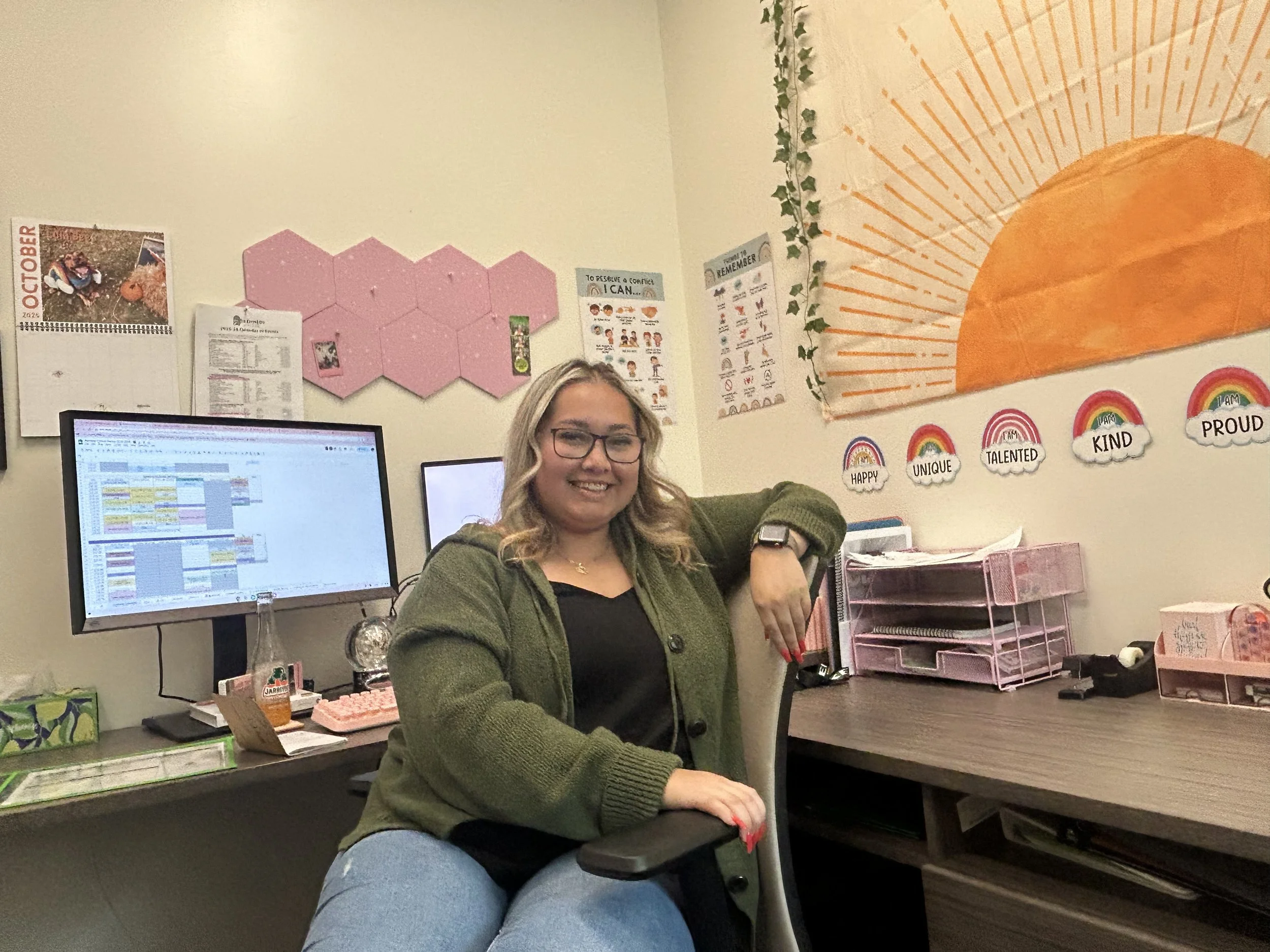 A woman sitting at a desk in a classroom or office, smiling at the camera, with a computer monitor and various office supplies around her. The classroom is decorated with a large wall tapestry, rainbow and cloud cutouts with positive words, and motivational posters.