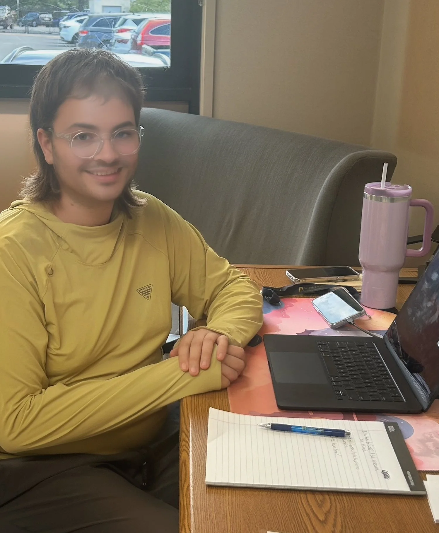 A person with short brown hair and glasses wearing a yellow hoodie, sitting at a table in a restaurant or cafe. There is a laptop, a notebook with a pen, and a pink tumbler on the table. Behind is a window showing parked cars outside.